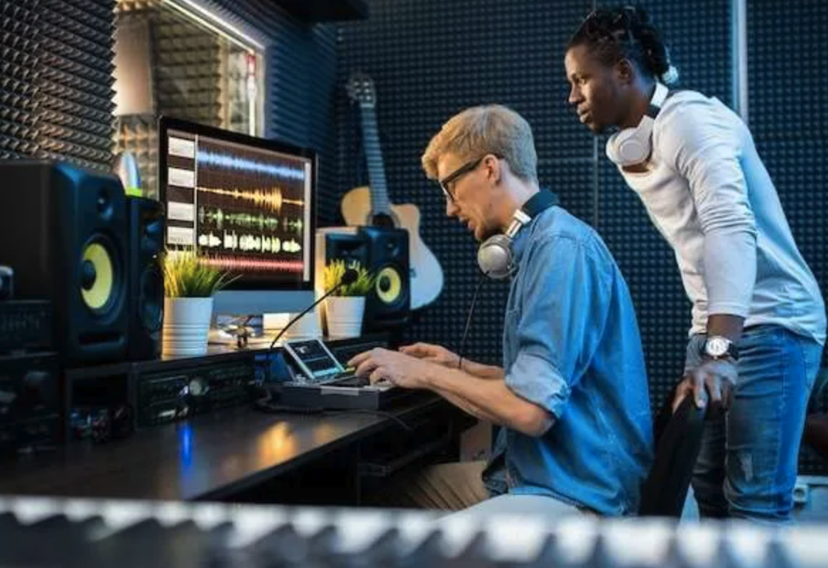 Two young men working in a recording studio. One is seated at a computer and audio equipment, wearing headphones and glasses, looking at the screen, while the other stands beside him, observing. The studio has soundproof foam walls, speakers, a guitar, and plants on the desk.