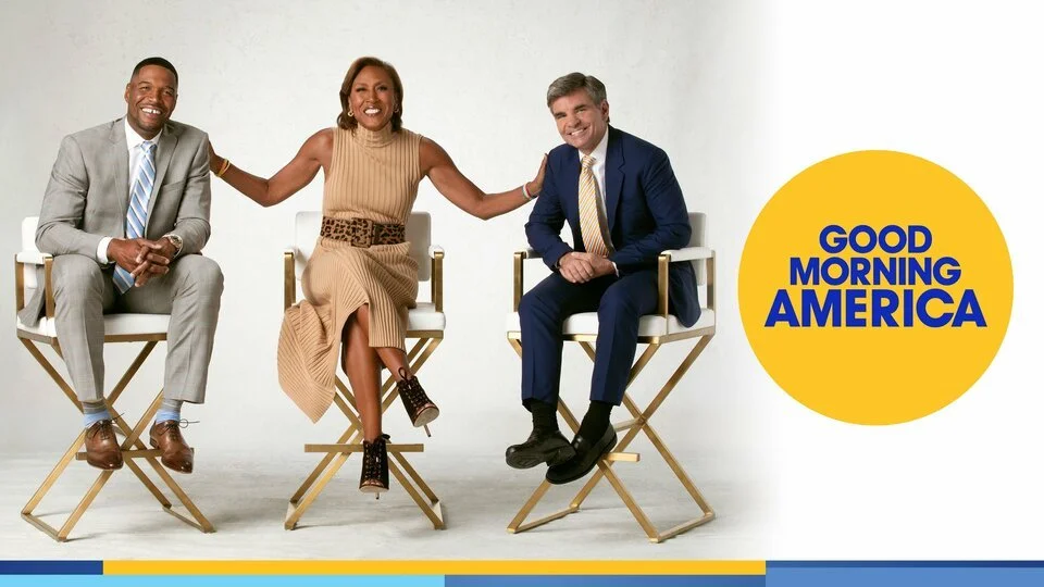 Three news anchors sitting in director's chairs, smiling, with a logo that says 'Good Morning America' on a yellow circle to their right.