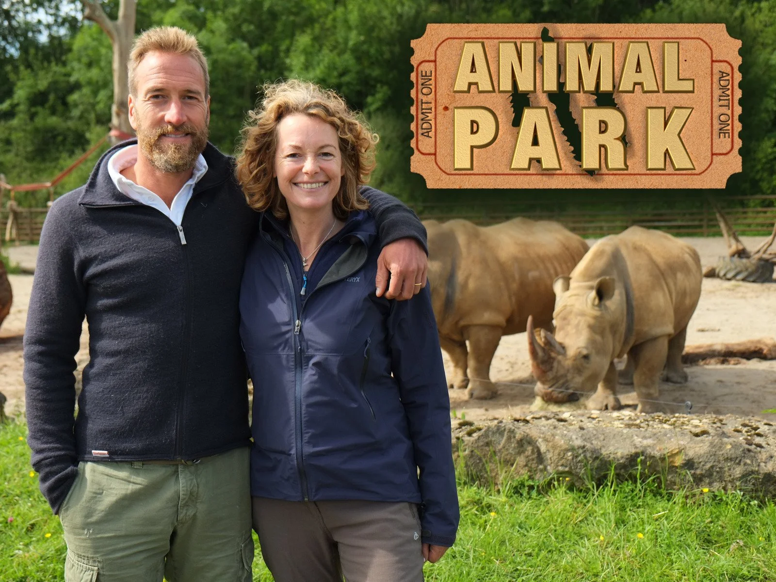 Smiling man and woman standing together outdoors at a zoo or animal park, with two rhinoceroses in the background and a sign that reads "Animal Park" in large letters.