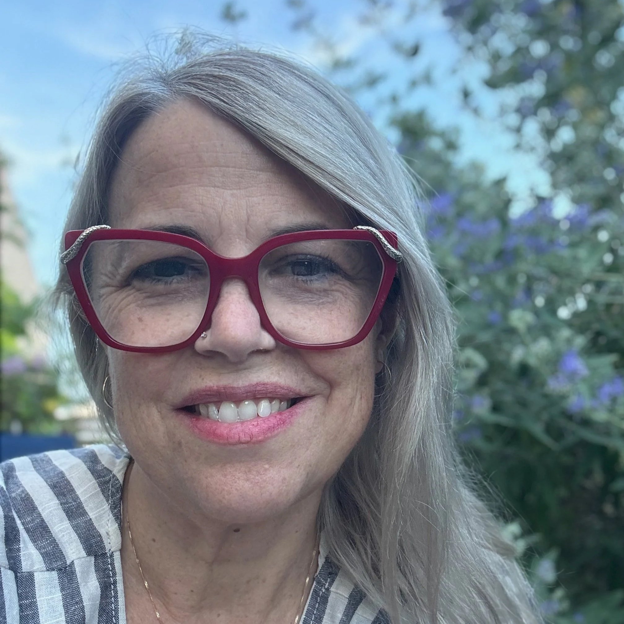 Kristin Mathis, a grey-blonde white woman with large red glasses looks directly at the camera.  She is standing in front of some periwinkle blue flowers with a blue sky behind her.