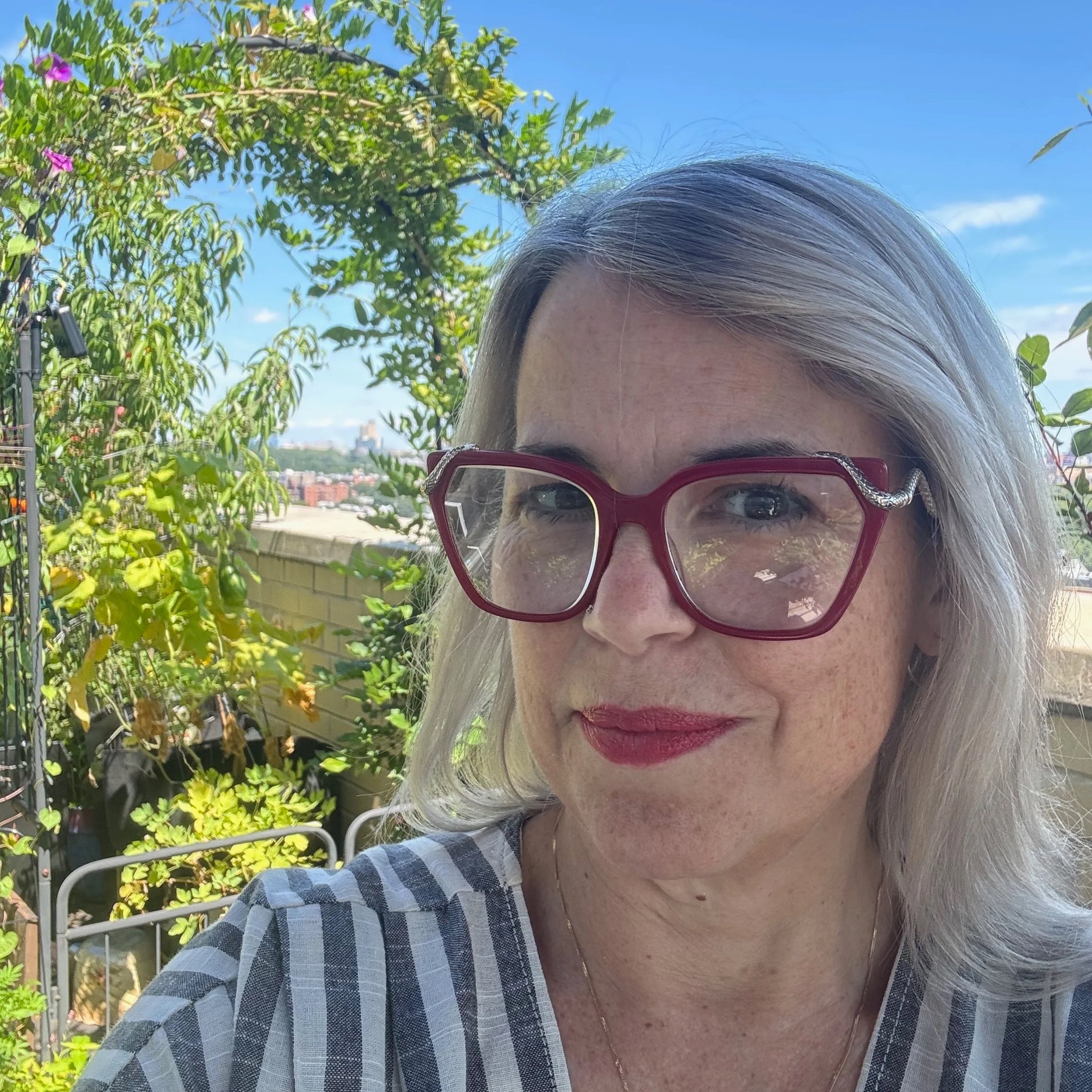 A white woman with large red glasses and grey-blonde hair wearing a striped shirt stands in front of a trellis covered with flowering vines.
