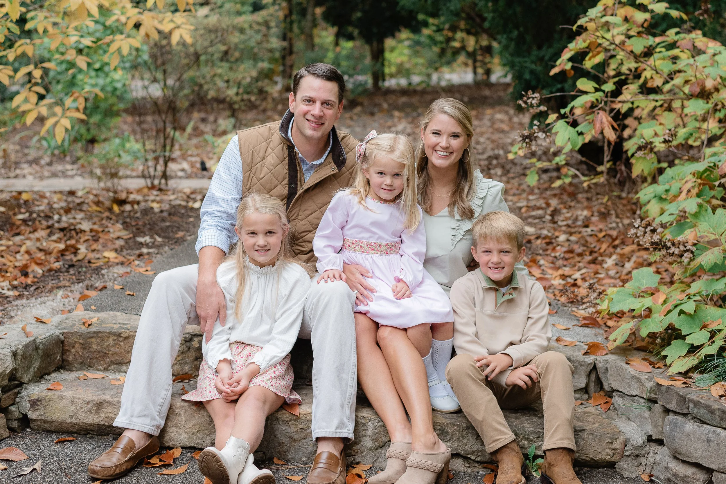 A family of six sitting on a stone staircase outdoors surrounded by autumn foliage, smiling for the camera.