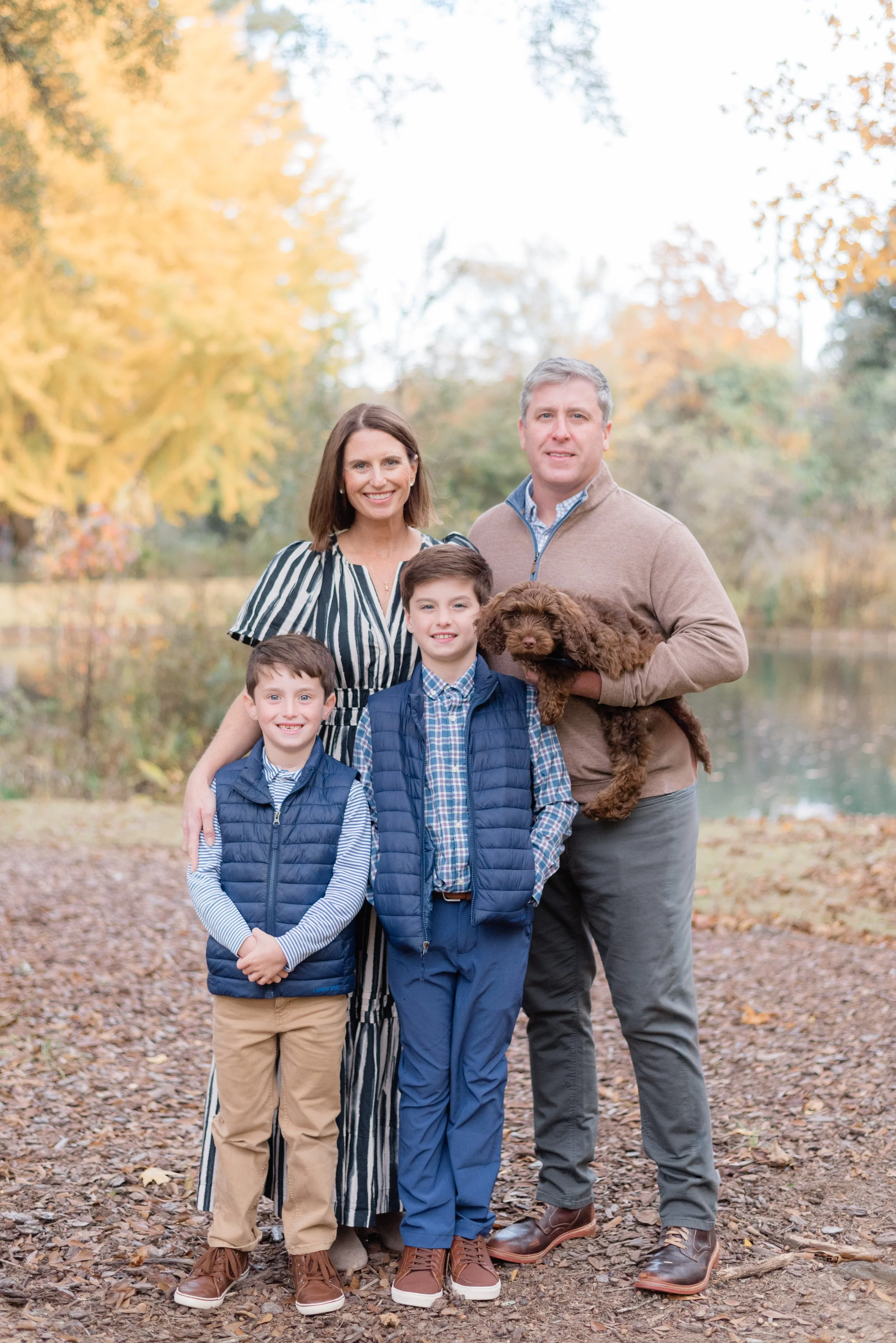 A family of four with a dog outdoors in autumn, smiling and posing for a photo on a leaf-covered path with yellow and orange trees in the background.