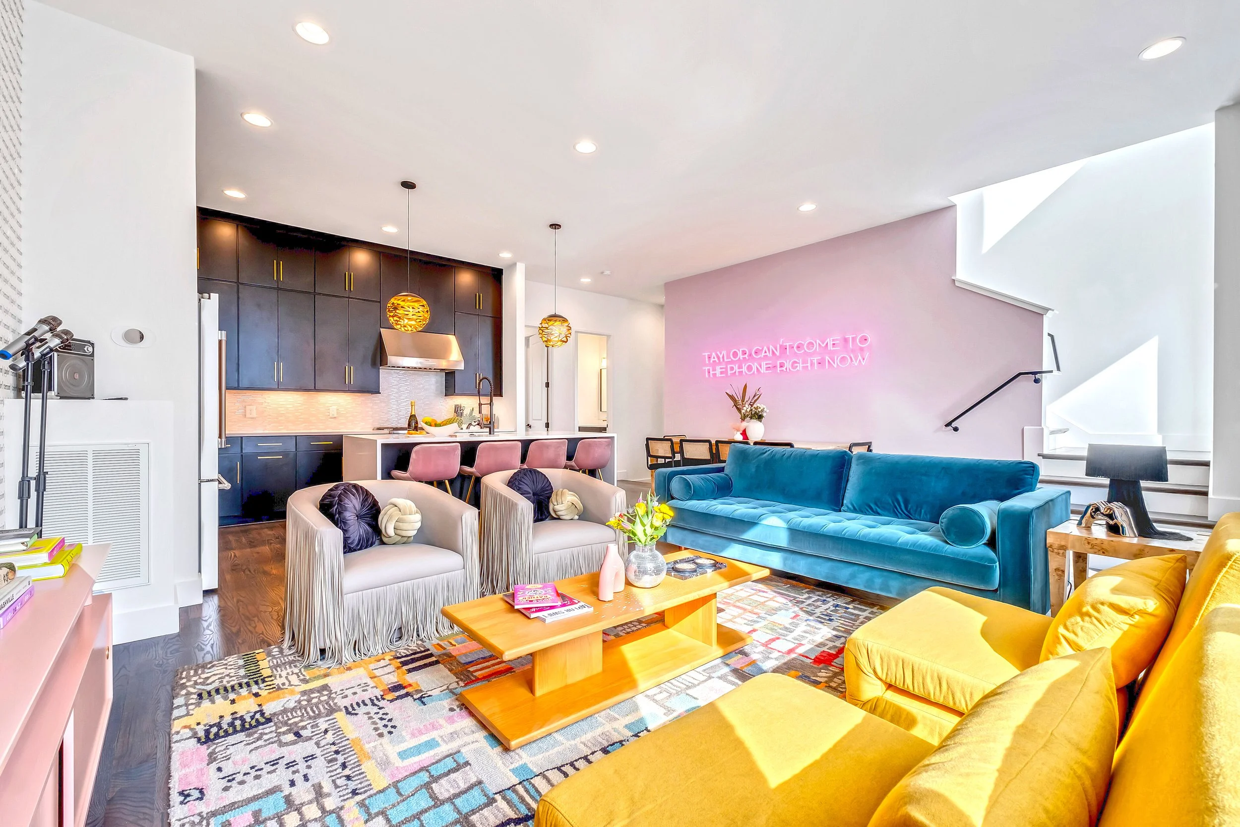 Interior view of a modern, colorful living room with a kitchen, featuring a blue velvet sofa, yellow chairs, pink and beige armchairs, a wooden coffee table with magazines and flowers, and a vibrant multicolored rug. The kitchen area has dark cabinet