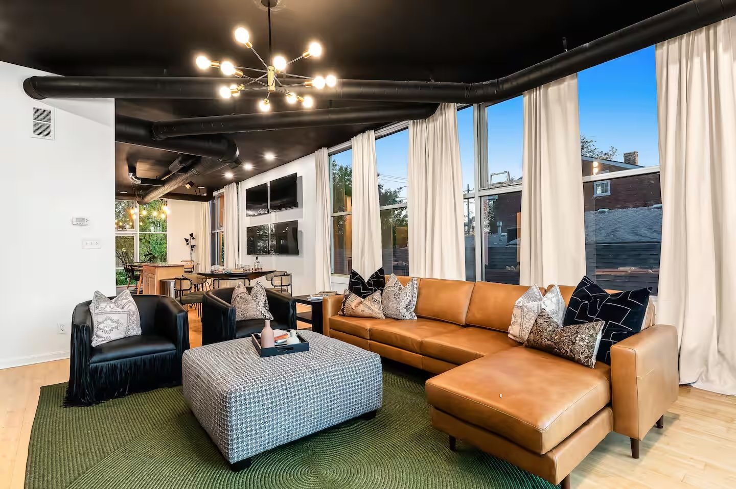 Modern living room with leather sectional sofa, black armchairs, patterned pillows, rectangular coffee table, green rug, large windows with white curtains, wall-mounted TVs, pendant lighting, and dining area in background.