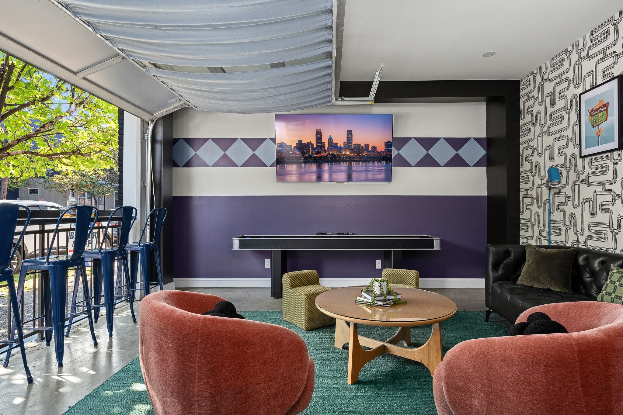 Indoor lounge area with pink velvet armchairs and a wooden coffee table, a black leather couch with pillows, a yellow armchair, and a green rug, with a wall-mounted TV showing a city skyline at sunset, and a patterned accent wall. Outdoors, a patio with chairs and a view of trees and parked cars.