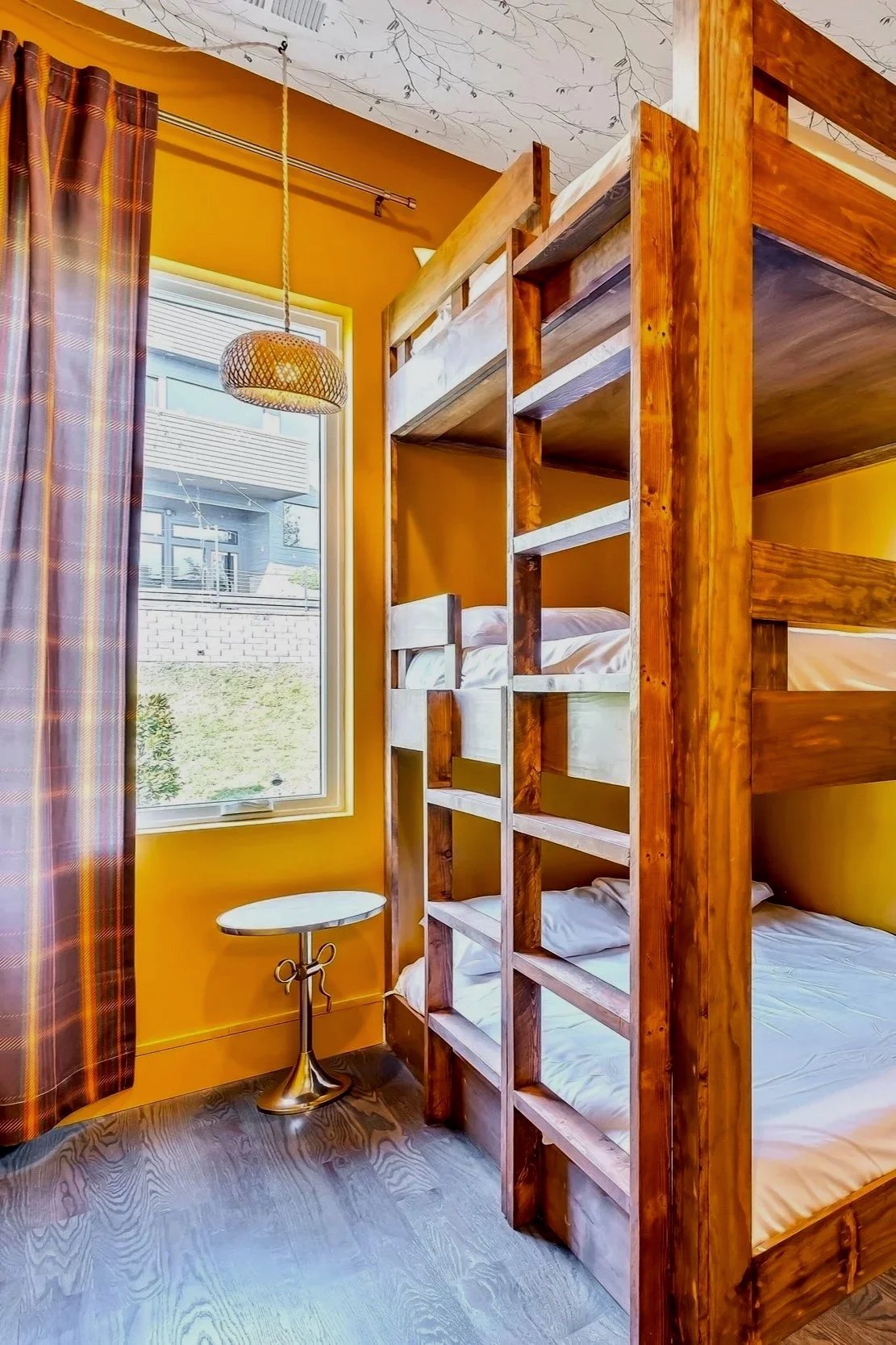A bedroom with yellow walls and a window with plaid curtains. There are wooden bunk beds and a small round side table with a metallic base next to the window.