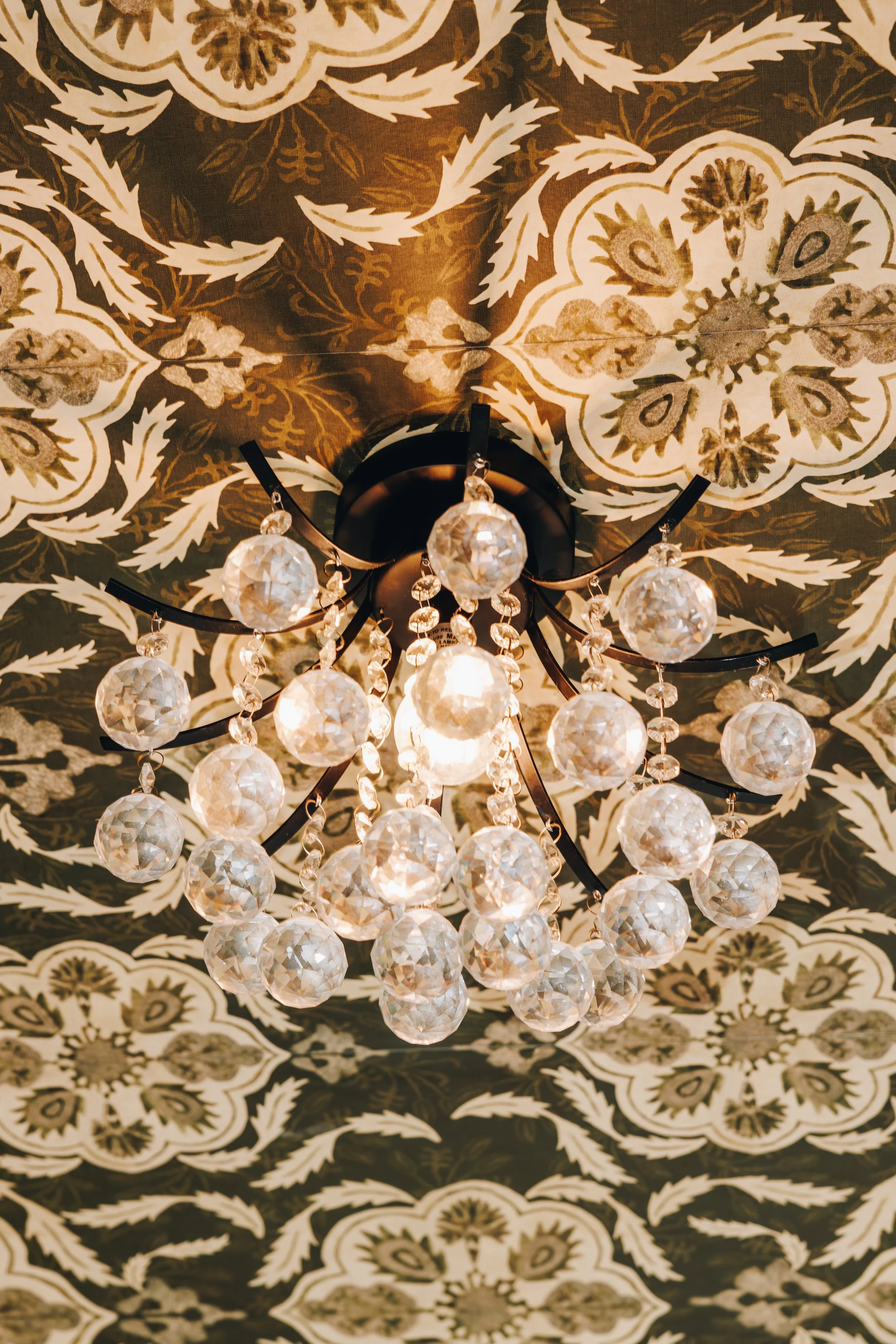 Close-up view of a chandelier with crystal ornaments hanging from a black metal frame, mounted on a ceiling with ornate patterned wallpaper in shades of brown, beige, and cream.