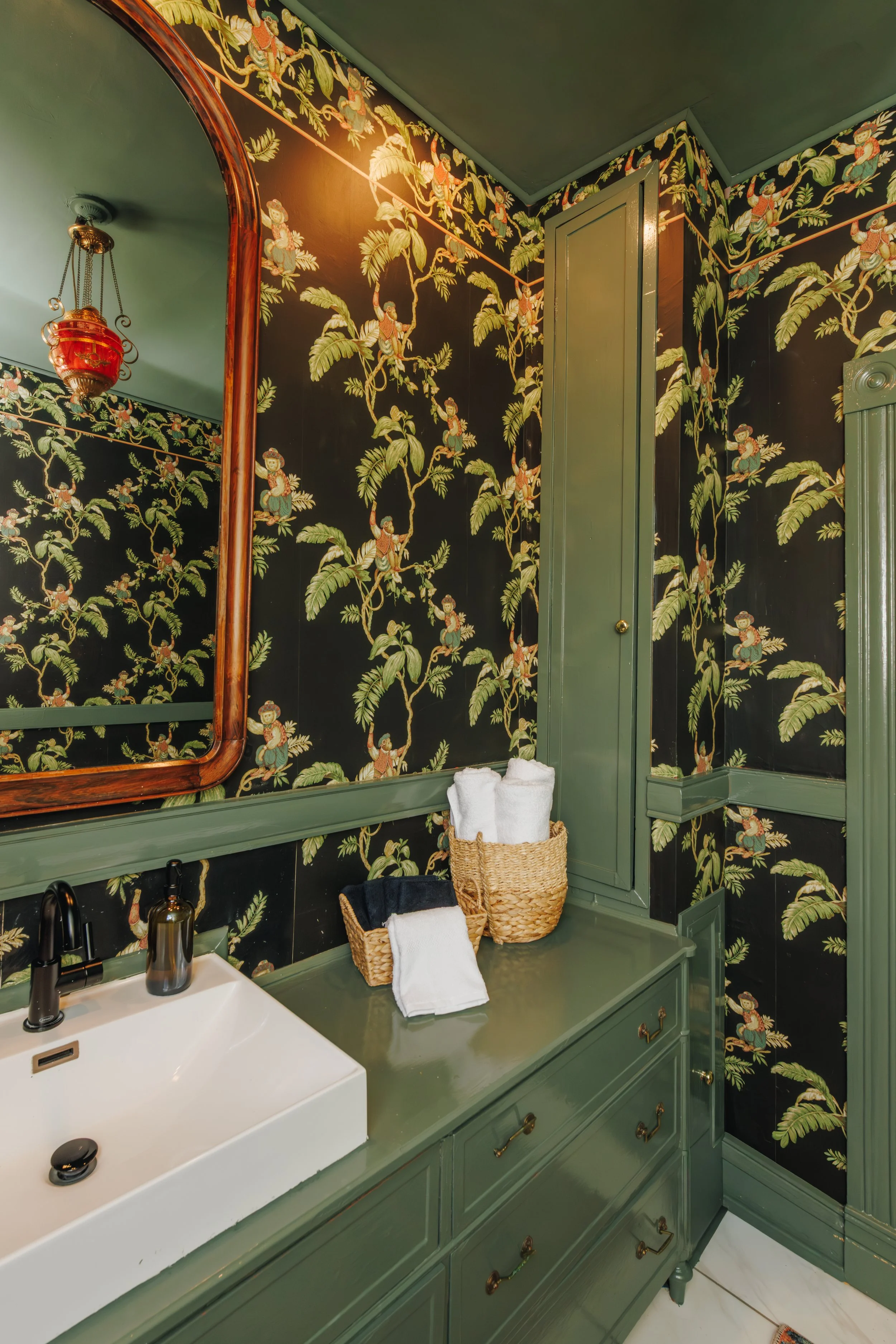Bathroom with green vintage-style vanity, black wallpaper with jungle plant and monkey design, mirror with wooden frame, red lantern light fixture, towels in baskets, and black faucet.