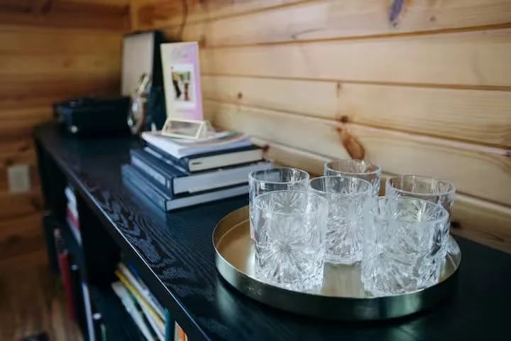 Six empty crystal glasses on a gold tray on a black table, with a stack of books, a framed picture, and a small black speaker in the background.