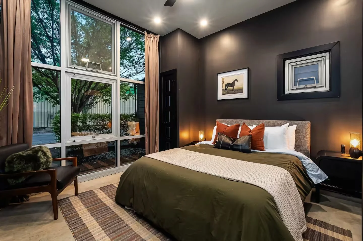 A bedroom with a large window showing trees outside, a bed with white and rust-colored pillows, dark gray walls, hanging framed pictures of a horse and a landscape, and a small wooden chair with a furry cushion.