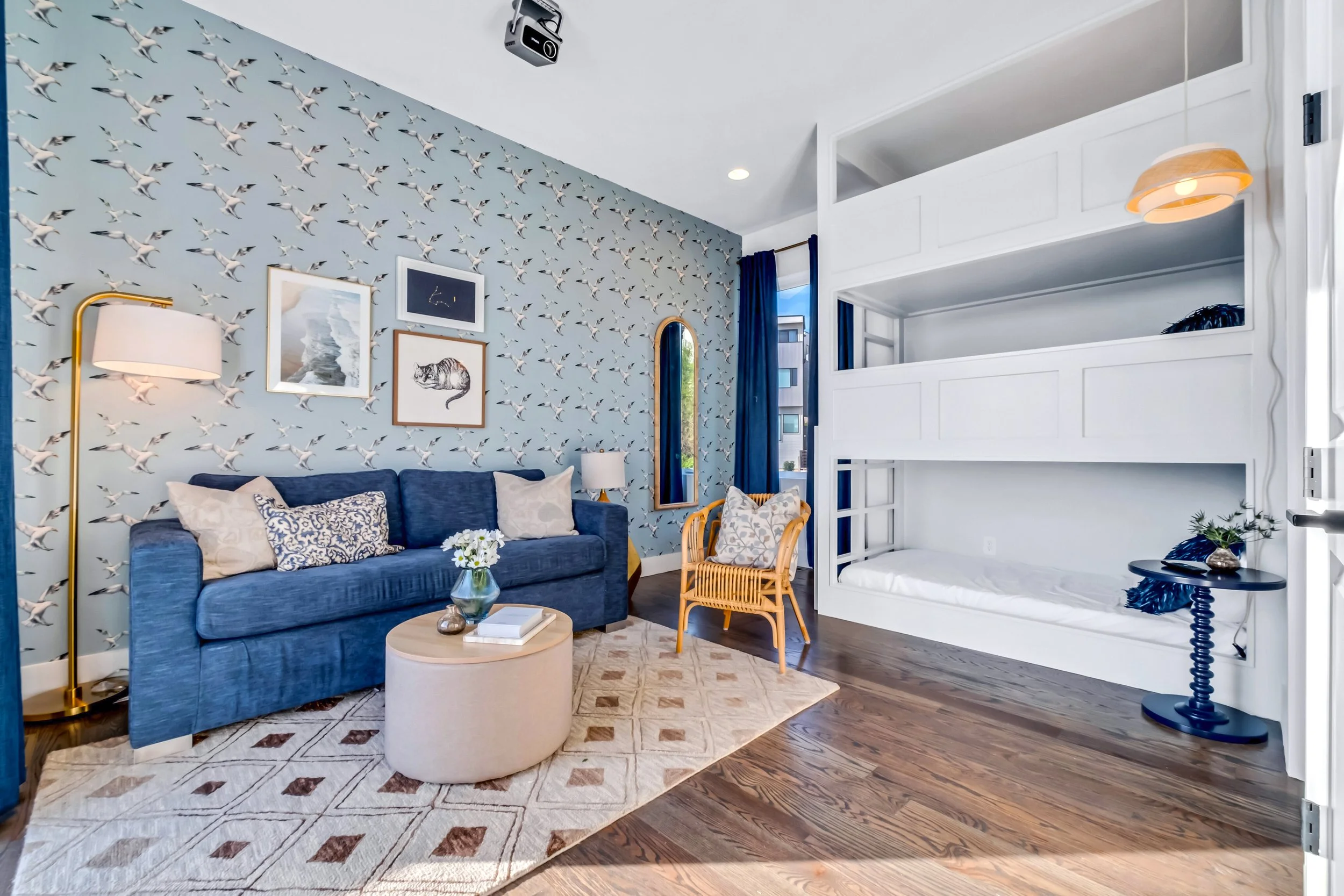 A living room with a blue sofa, patterned pillows, a round beige ottoman, a modern wall with seagull wallpaper, tropical artwork, and a bunk bed with white framing in the corner.