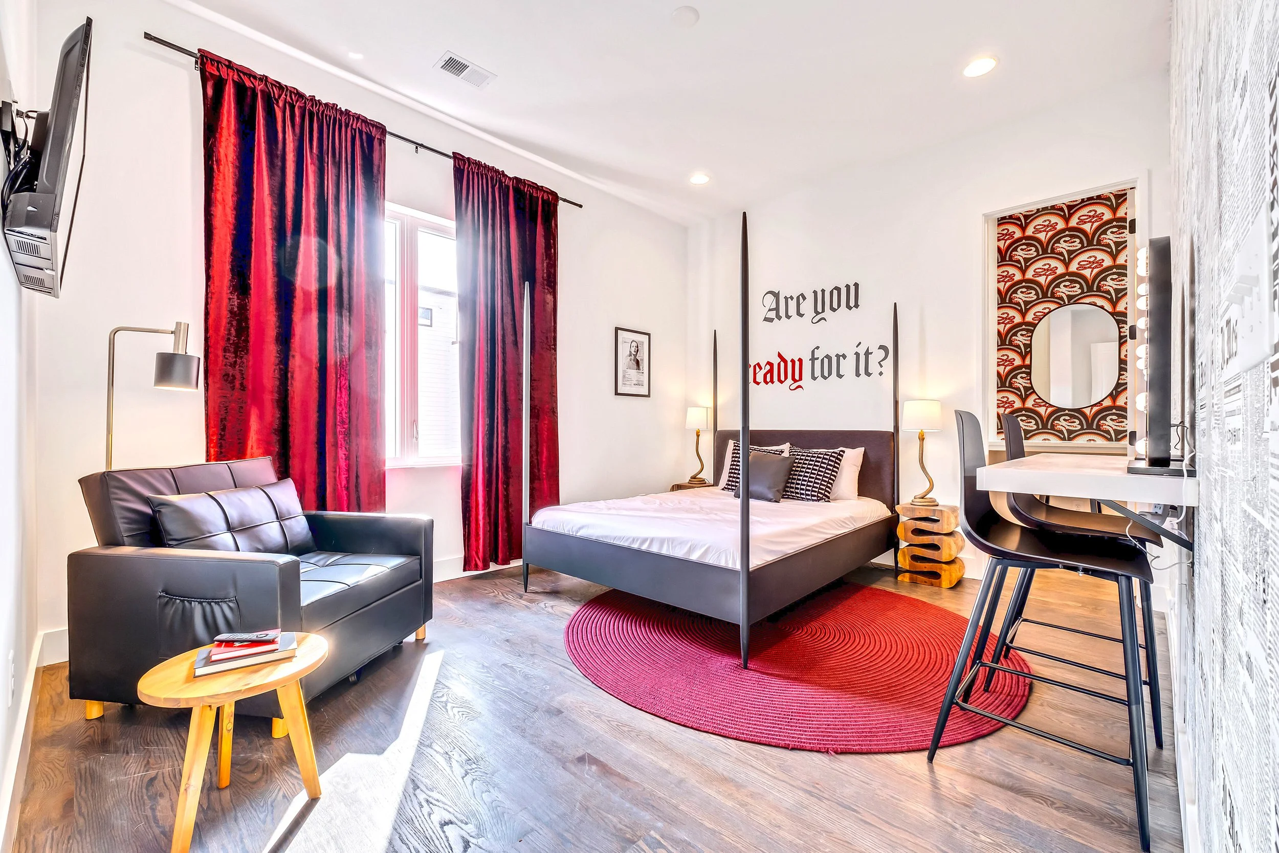 A modern bedroom with a bed, black headboard, and white bedding, flanked by two lamps. Red curtains cover a window, and a red circular rug is on the wooden floor. There's a black leather armchair with pillows, a small wooden side table with magazines