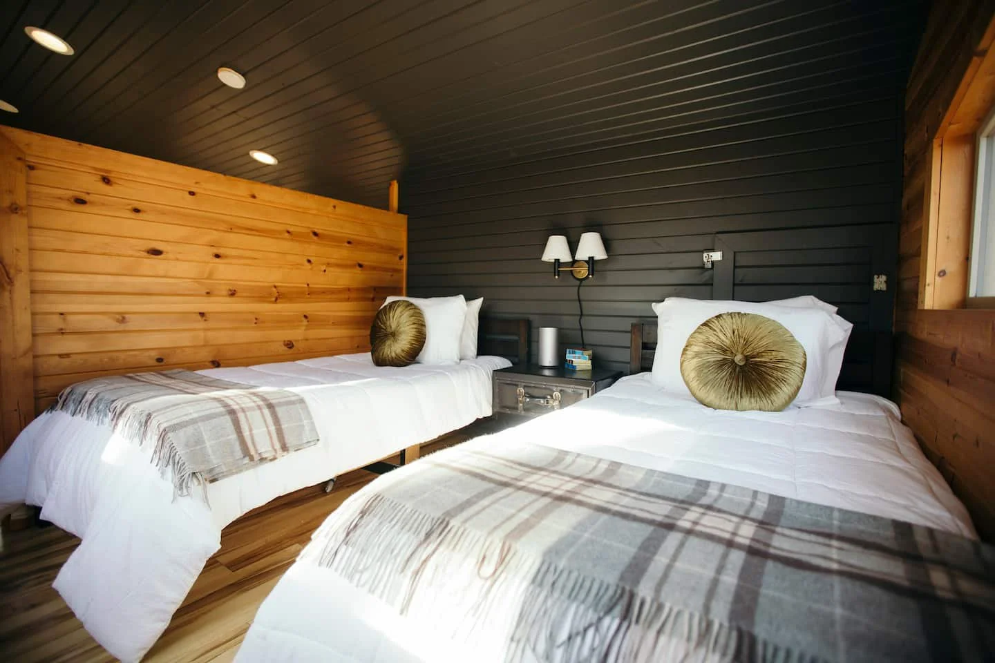 Two beds in a cozy bedroom with wood paneling, each with white bedding, plaid throws, and round decorative pillows. There is a nightstand with a lamp and items between the beds, and a window on the right side.