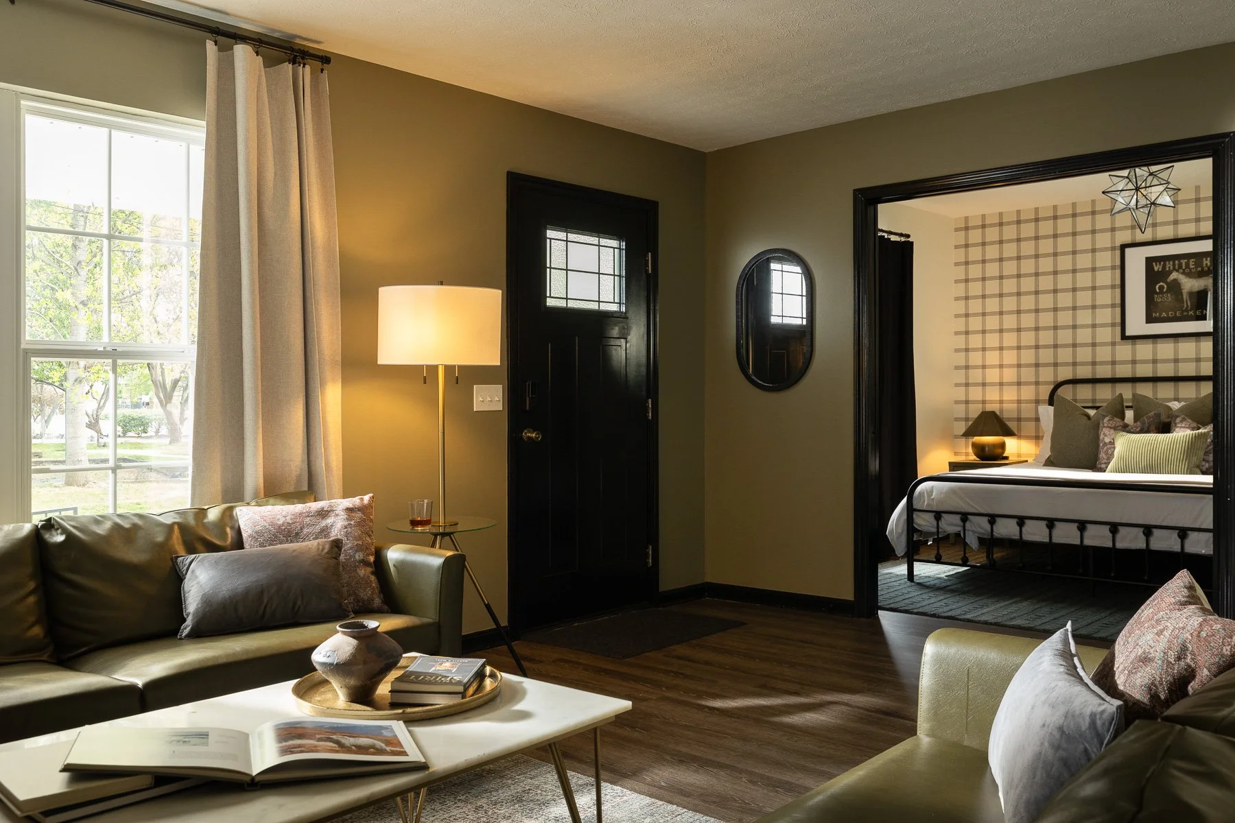 Living room with a large window, beige curtains, a beige sofa with pillows, a coffee table with magazines, and a floor lamp. Adjacent to the living room is a bedroom with a bed, side tables with lamps, and a wall with plaid wallpaper.