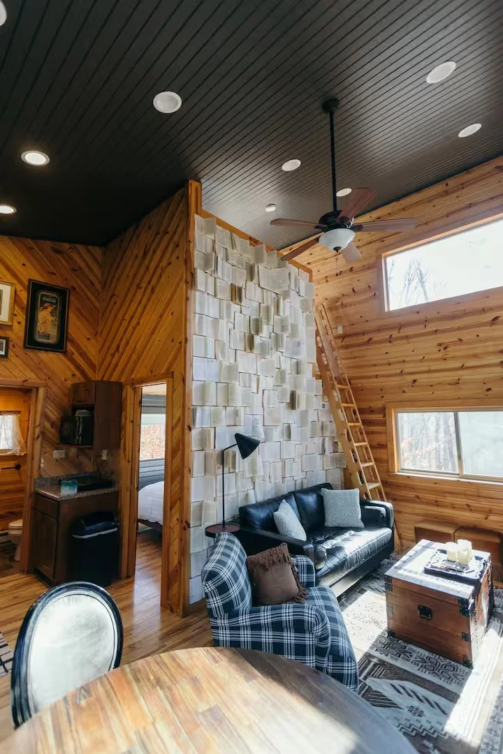 Living room with wooden walls and ceiling, featuring a black leather sofa, checked armchair, wooden coffee table, and large windows. There is a stone accent wall with a ceiling fan and a ladder leading to an upper level.