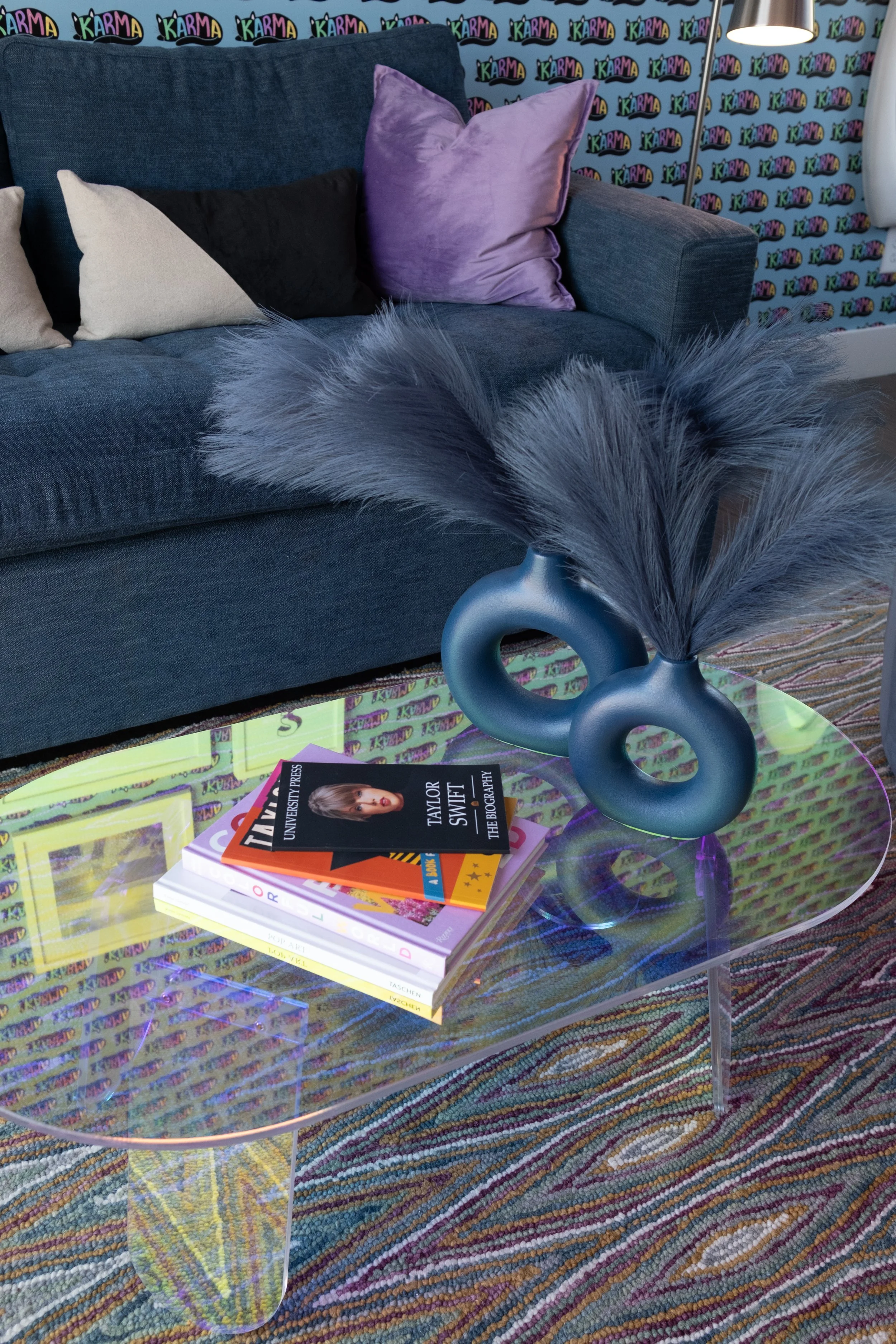 A dark blue fabric sofa with four throw pillows in beige, black, and purple. A black and purple wallpaper with the word 'KARMA' repeated repeatedly in colorful fonts. A round transparent glass table with a colorful, iridescent surface and a decorative black ceramic vase with tall gray feathers. The table also holds books, including one titled 'Taylor Swift' and a magazine. A tall, thin standing lamp with a silver pole and a black lampshade is visible to the right. The setup is indoors, on a multicolored striped rug.