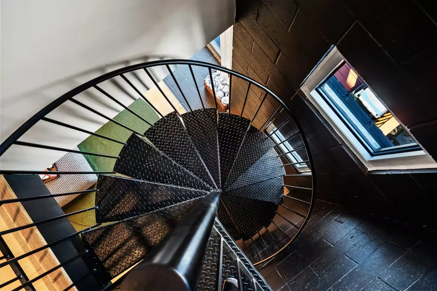 A black spiral staircase with black metal railings and diamond-patterned steps, viewed from above near a window that shows the outside street.
