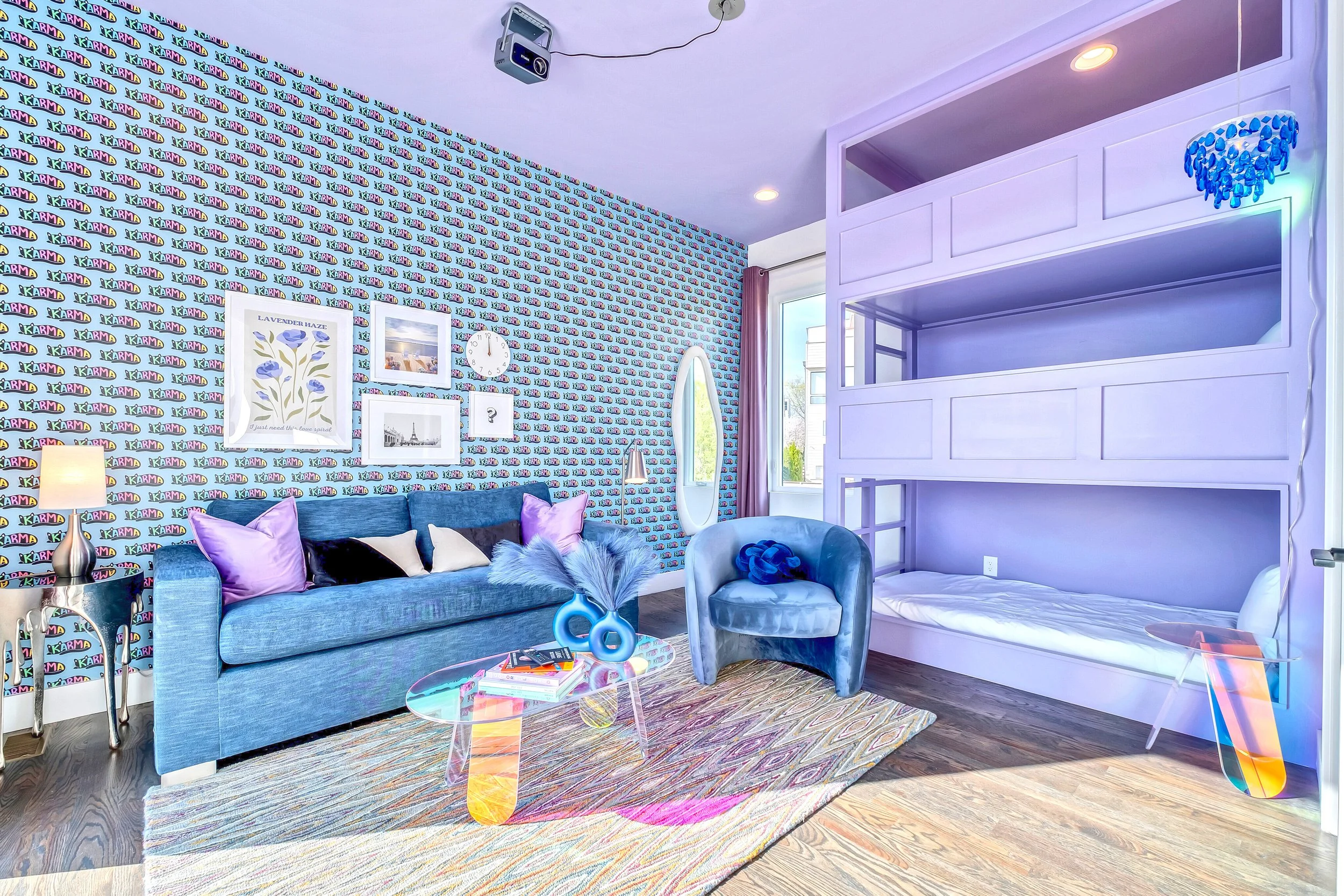 Living room with blue velvet sofa, purple and black pillows, wall with repeating 'Karma' pattern, framed art, round mirror, a modern lamp, a clear glass coffee table with books, colorful rug, and a white bunk bed with blue chandelier, near a window w
