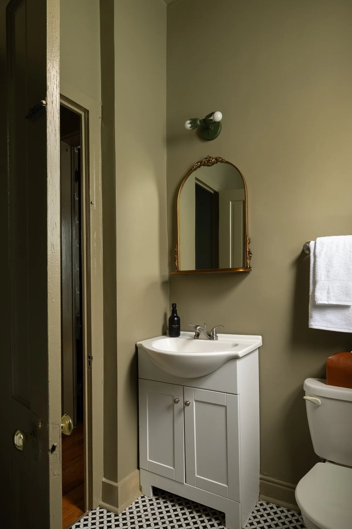 Small bathroom with a white sink and cabinet, a mirror with a gold frame, a towel rack with white towel, and a black soap dispenser on the sink. The floor has black and white mosaic tiles. There is a wall sconce with two bulbs above the mirror, and a door on the left side of the image.