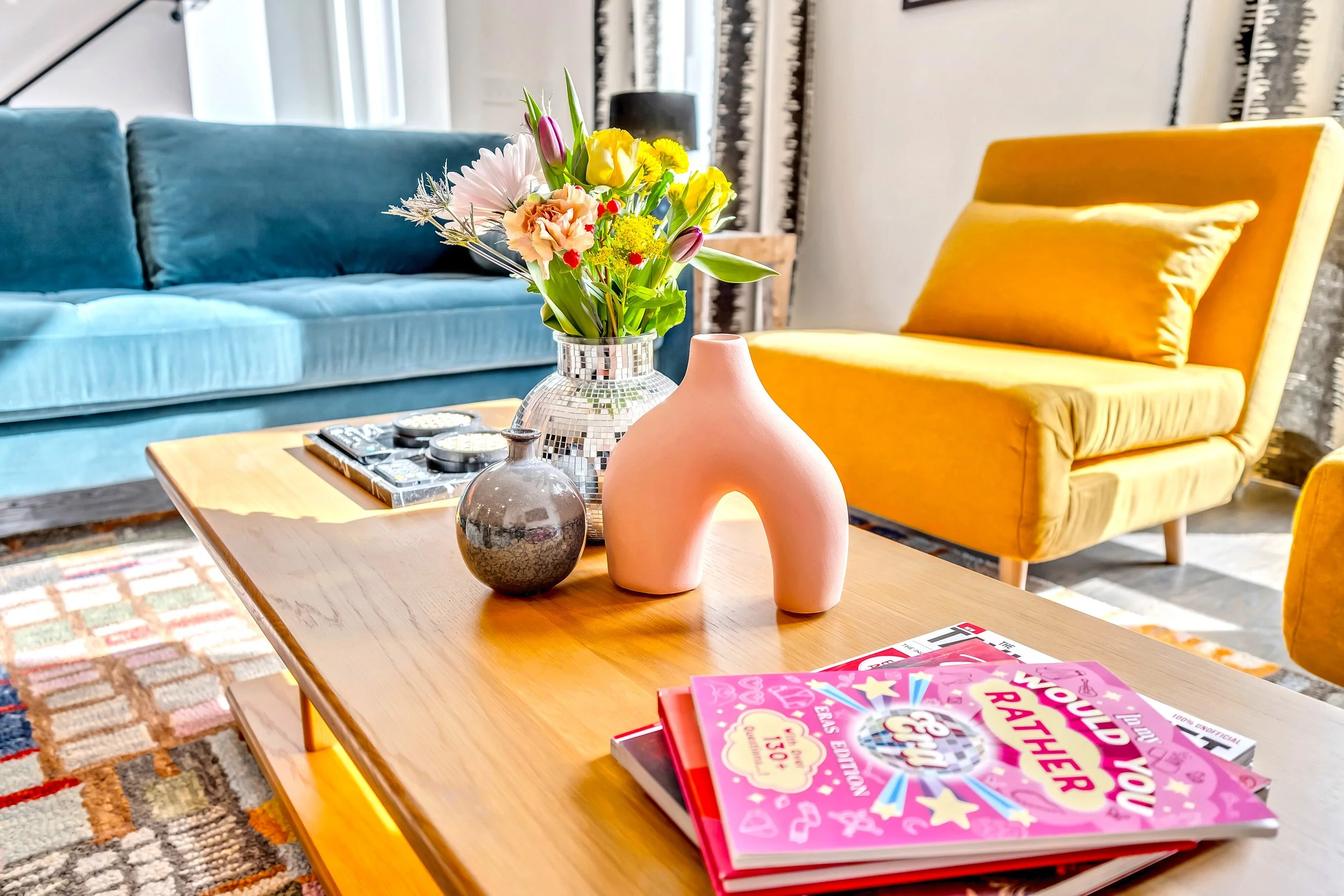 A living room with a wooden coffee table holding a bouquet of colorful flowers in a silver vase, complemented by pink and gray decorative vases, and surrounded by a blue sofa and a yellow armchair. Magazines are on the table and framed pictures hang 