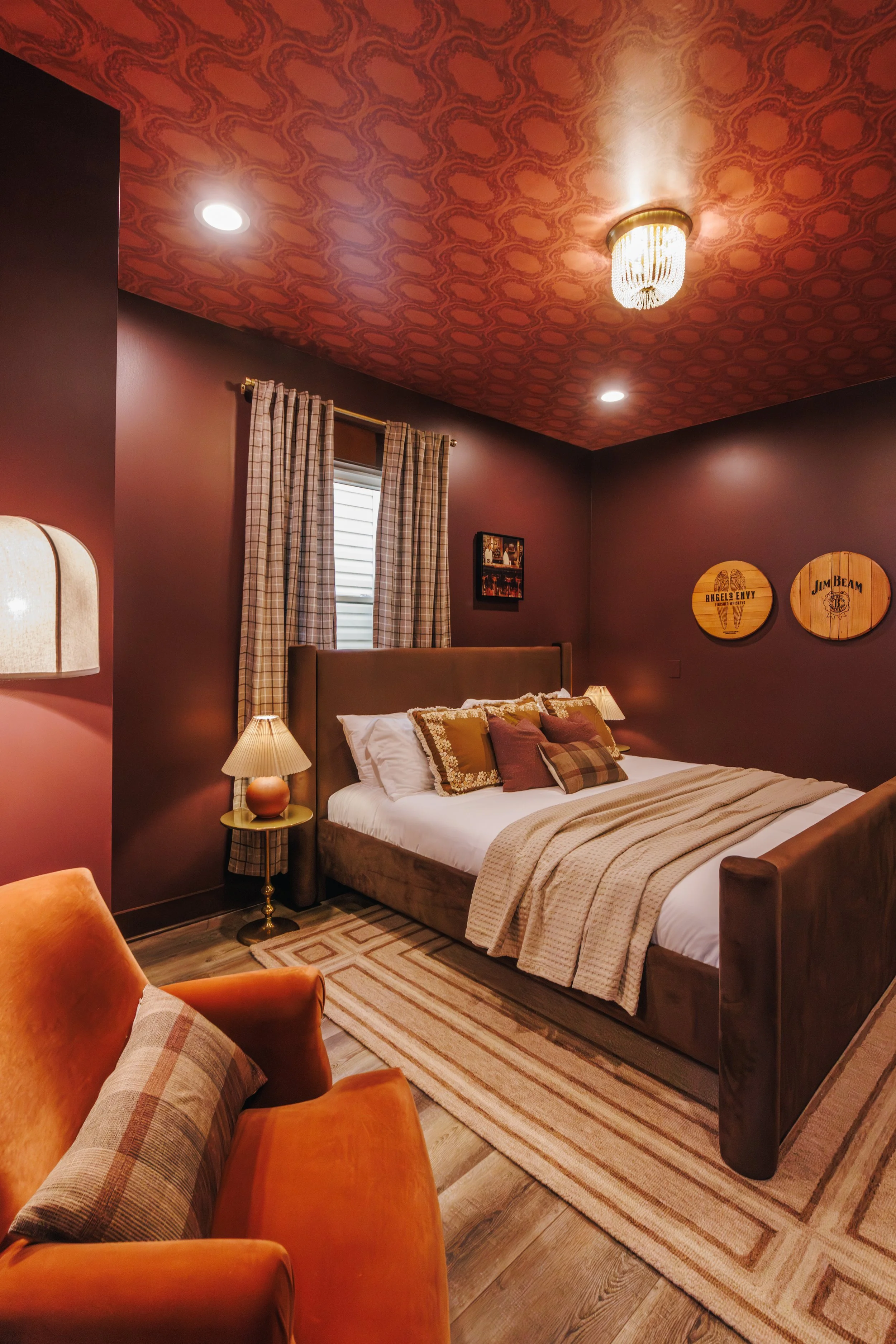 A cozy bedroom with a large bed, pillows, a beige blanket, and a wooden headboard. There is a small round side table with a lamp on it. The walls are painted a warm dark color, and there are plaid curtains over a window. Two circular wall decorations hang on the wall, one with the text "Angels Envy" and the other with the text "Jim Beam." The ceiling is decorated with patterned wallpaper and a chandelier, with additional recessed lighting.