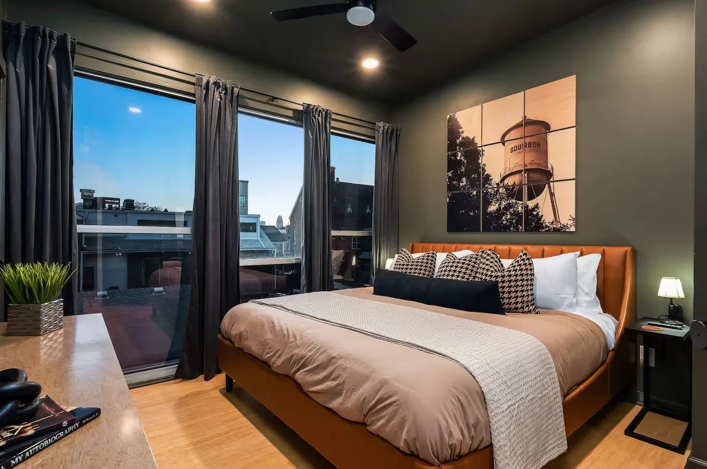 A modern bedroom with a large bed, tan headboard, patterned and white pillows, beige bedding, and a nightstand with a small lamp. There is a large window with dark curtains, a green plant on the desk, and a multi-panel wall art depicting a water towe