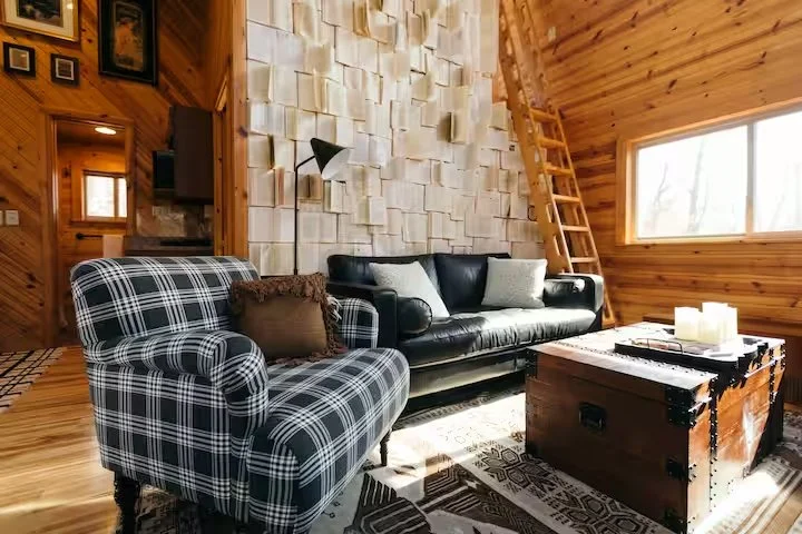 Living room with plaid armchair, black leather sofa, wooden walls, window, ladder, wood paneling, and decorative wall panels.