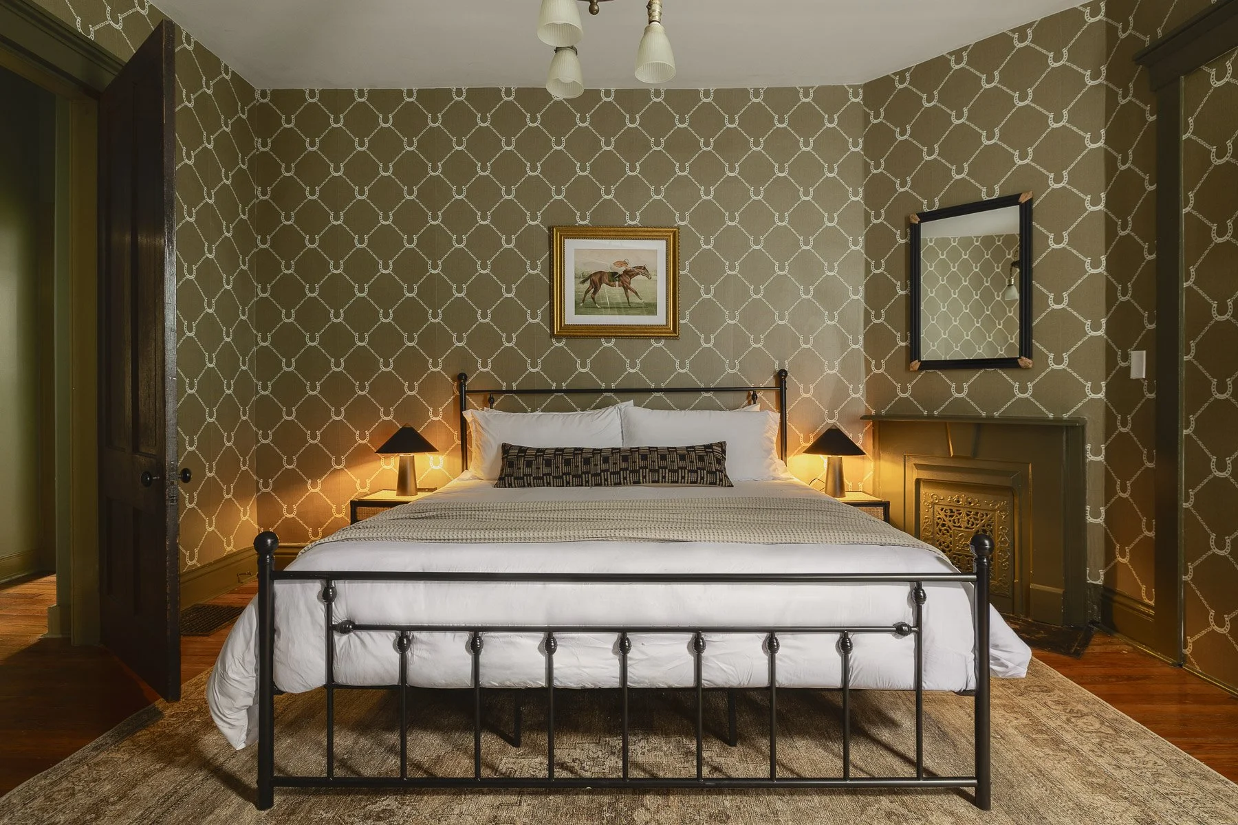 A bedroom with a black metal bed frame, white bedding, and pillows. Two bedside lamps with black shades are on either side of the bed. The wall behind has a patterned wallpaper with a chain-link design, and a framed picture of a horse and rider hangs above the bed. A mirror is on the right wall, reflecting part of the room, and a decorative fireplace insert is on the right wall. The ceiling has a ceiling fan with light fixtures.