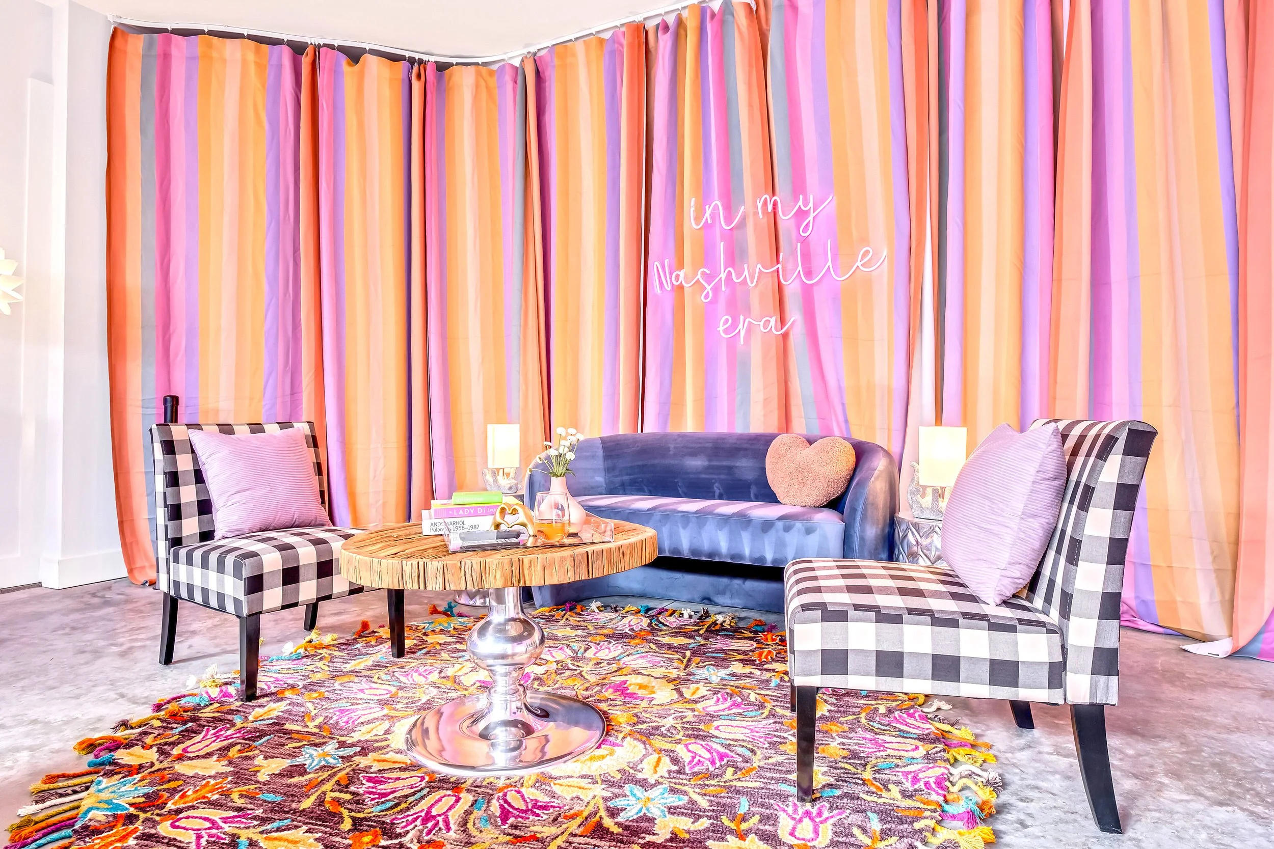 Colorful room with striped curtains, neon sign reading "in my Nashville era," a purple velvet sofa with pillows, checkered chairs, a floral area rug, a round coffee table with books and drinks, and decorative lamps.