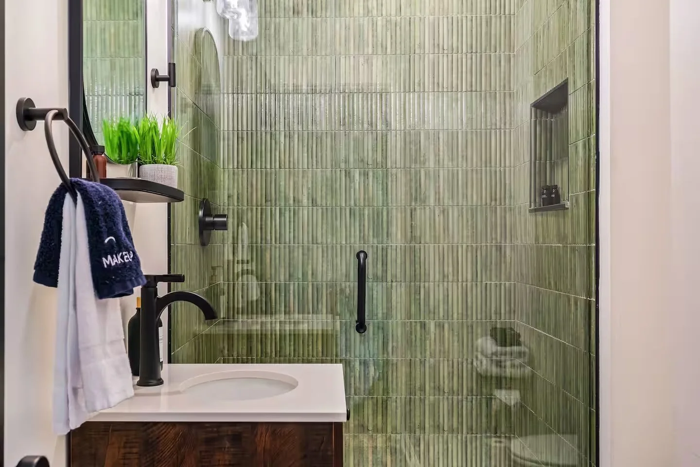 Bathroom with a green-tiled walk-in shower, black fixtures, and a small sink with a marble countertop. A towel ring with towels and a shelf with a plant are visible on the left side.