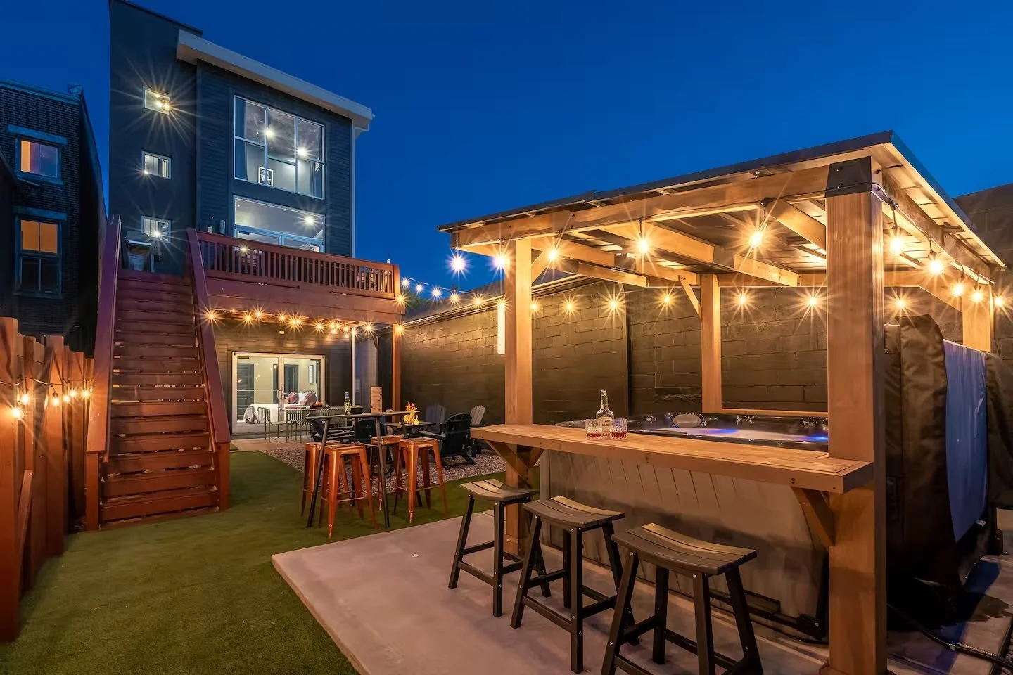 Nighttime outdoor backyard with string lights, a hot tub under a wooden shelter, a high bar table with stools, and a patio area with chairs and a table.