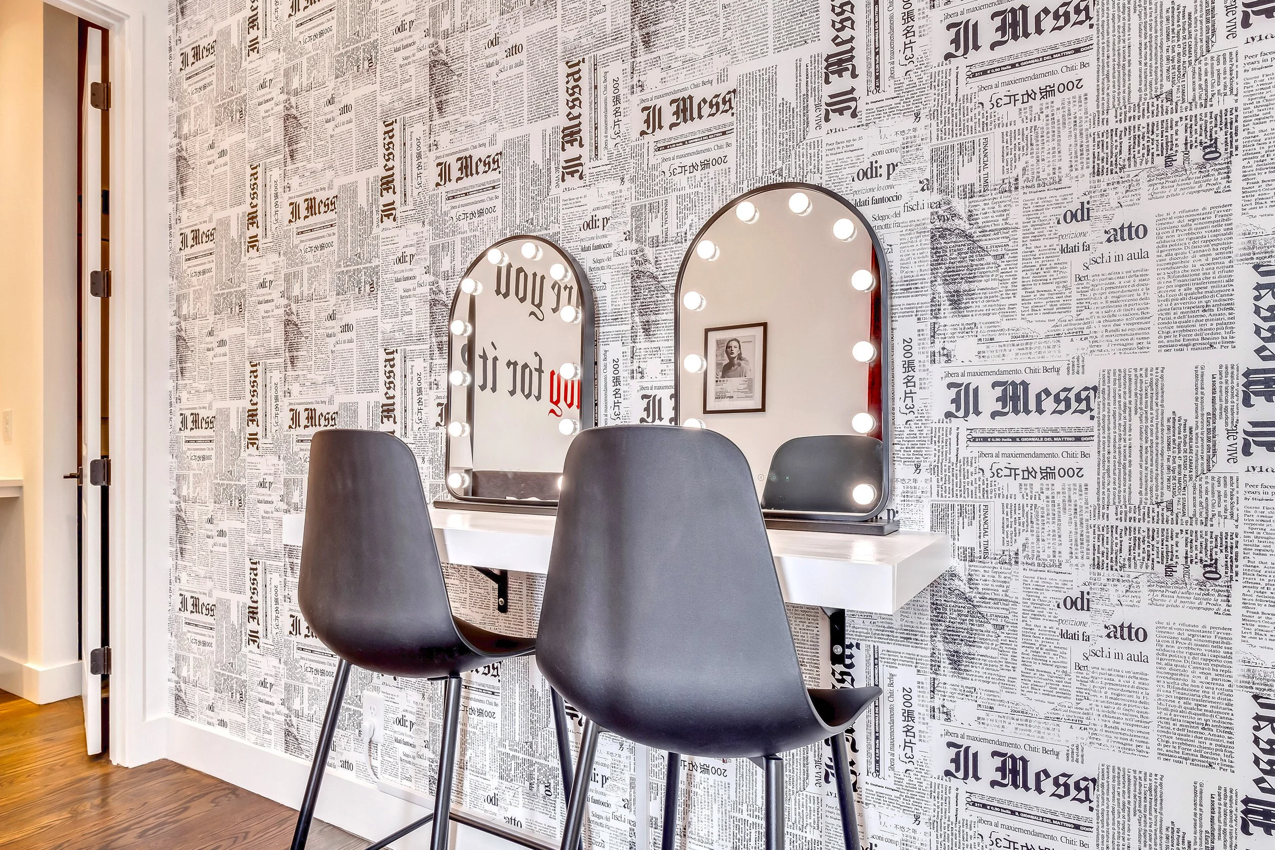 A stylish dressing area with two black chairs in front of a wall covered in newspaper print wallpaper, two mirrors with light bulbs, and a framed photograph.