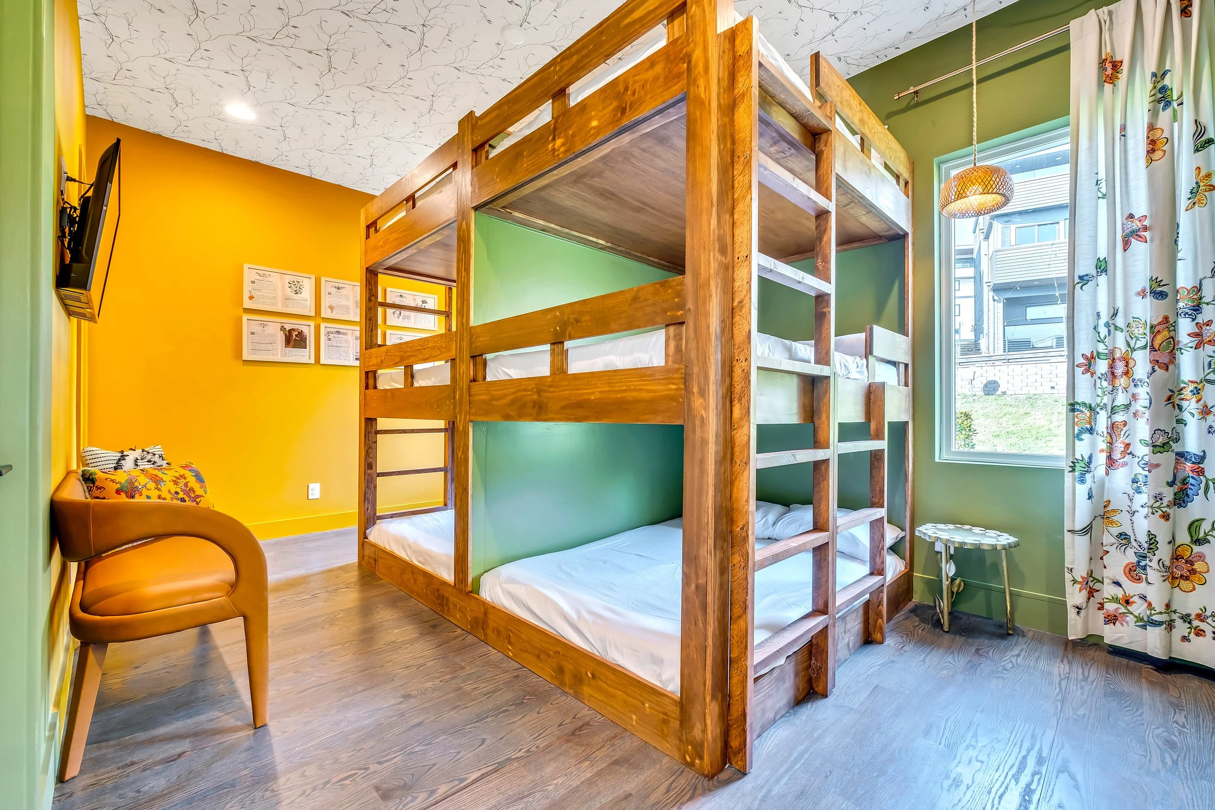Interior of a colorful room with a wooden bunk bed, yellow and green walls, a window with butterfly-patterned curtains, a tan armchair, and decor on the walls.