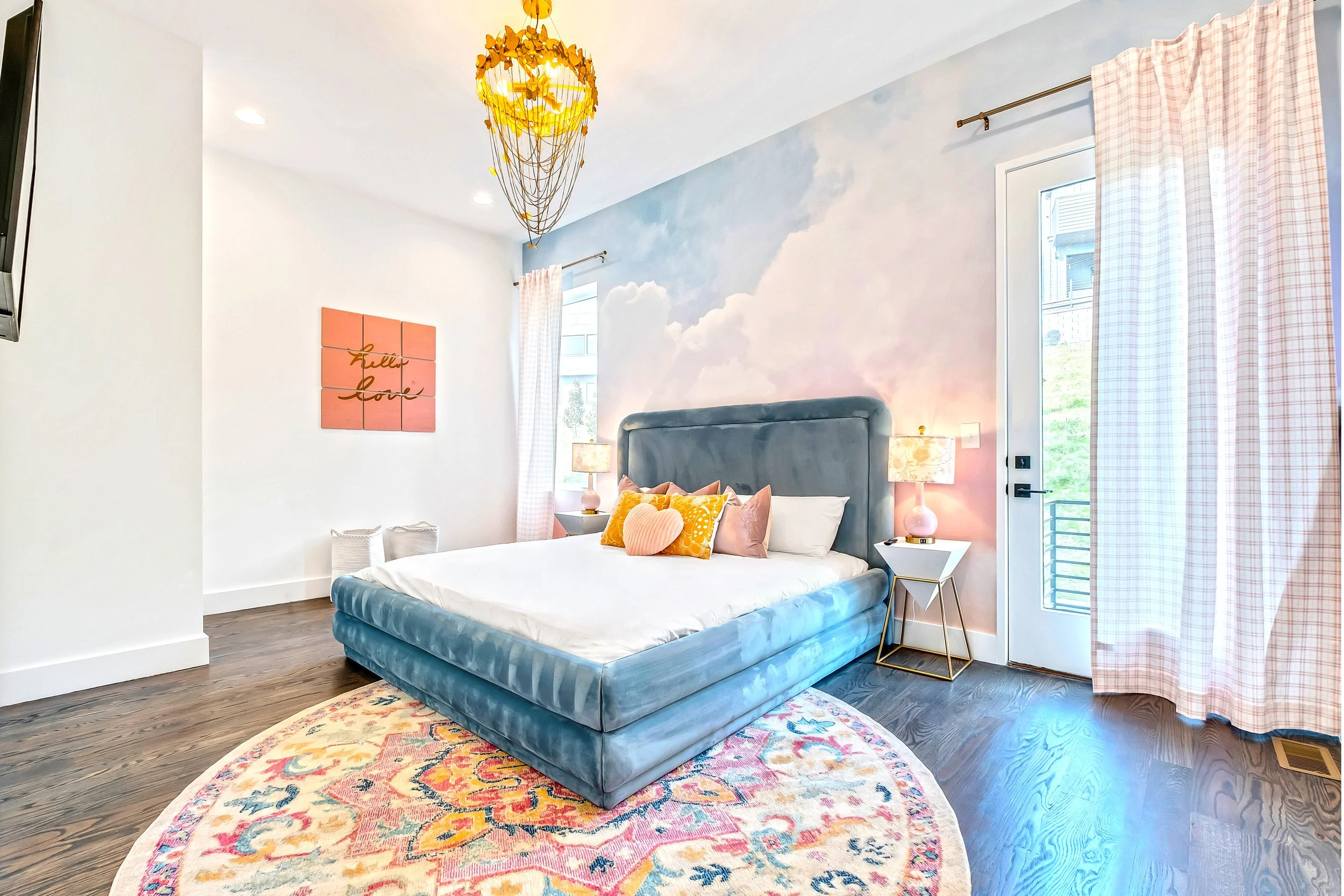 A bedroom with a sky-themed mural wall, a bed with colorful pillows, a patterned rug, and pink curtains near a door with a window.