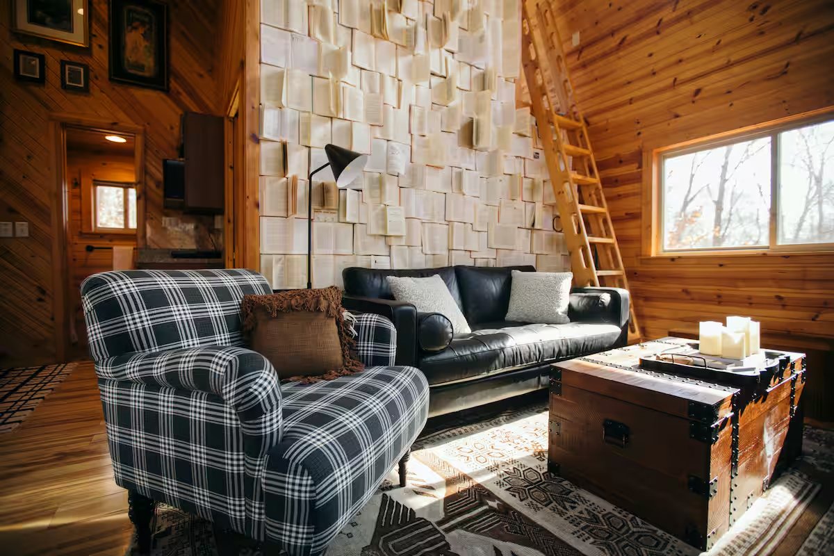 Cozy living room with wood-paneled walls, a black leather sofa and a black and white plaid armchair, a wooden trunk coffee table, and a unique wall with stacked books as decoration. A ladder leads to a loft, and sunny windows illuminate the space.