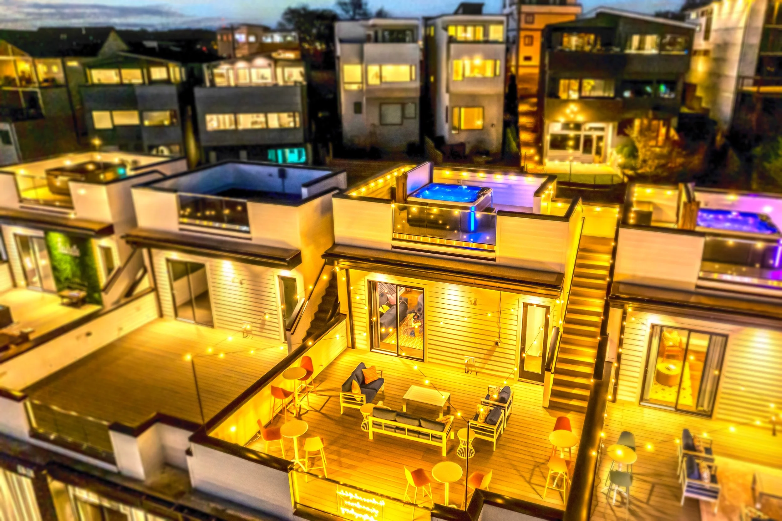 Aerial view of modern multi-story apartment buildings at night illuminated with warm yellow and purple string lights, with a terrace and hot tub visible on the rooftop of one building.