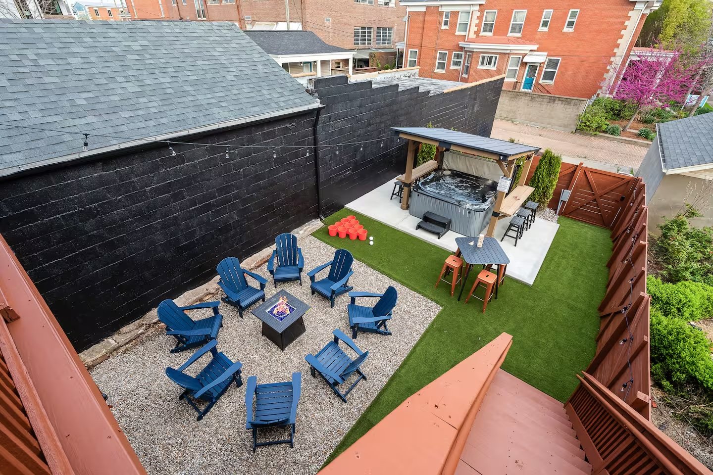 A rooftop backyard with blue Adirondack chairs around a fire pit, a hot tub under a wooden pergola, and a fenced grassy area with a high fence and surrounding trees.