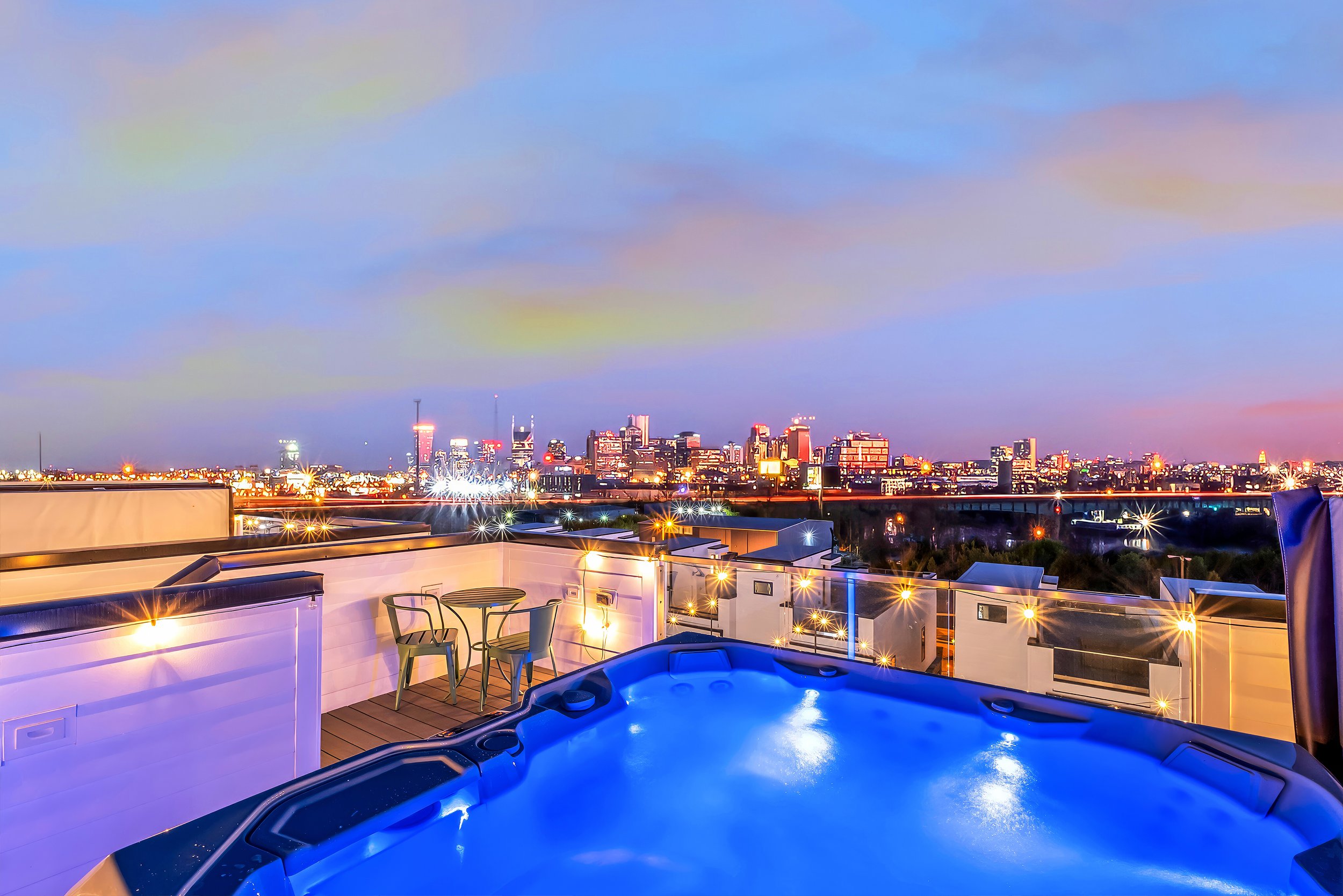 City skyline at dusk viewed from a rooftop balcony with a hot tub and outdoor furniture, including a table and chairs.