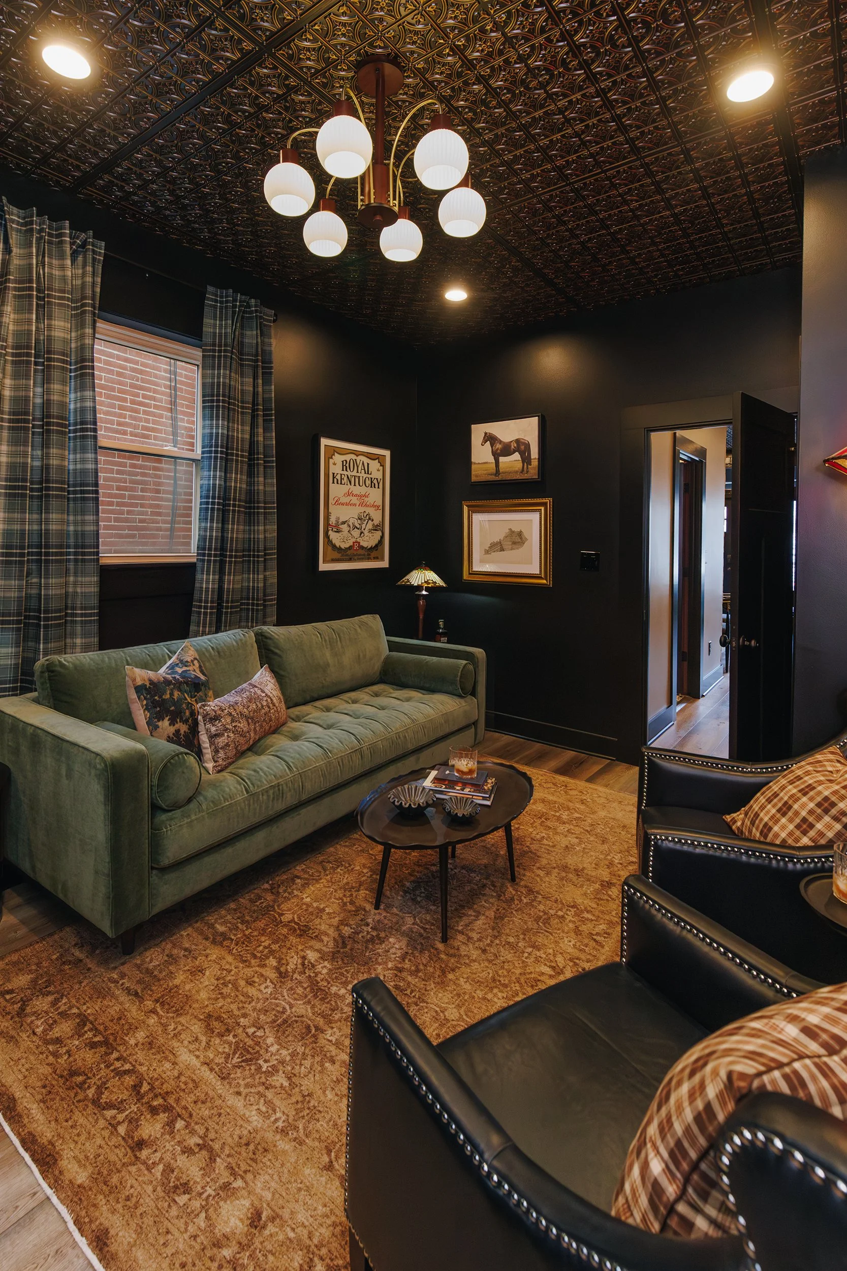 Living room with dark walls, plaid curtains, green velvet sofa with pillows, black leather chairs, a round coffee table, framed pictures on the wall, a lamp with a stained glass shade, and a decorative ceiling with warm lighting.