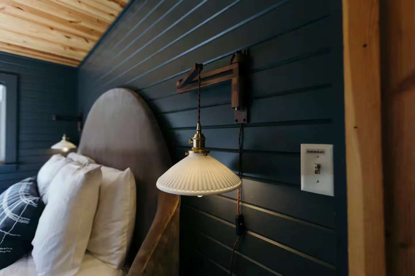 Close-up of a bedroom headboard with a wall-mounted lamp, electrical outlet labeled 'OVERHEAD,' and a bed with pillows.