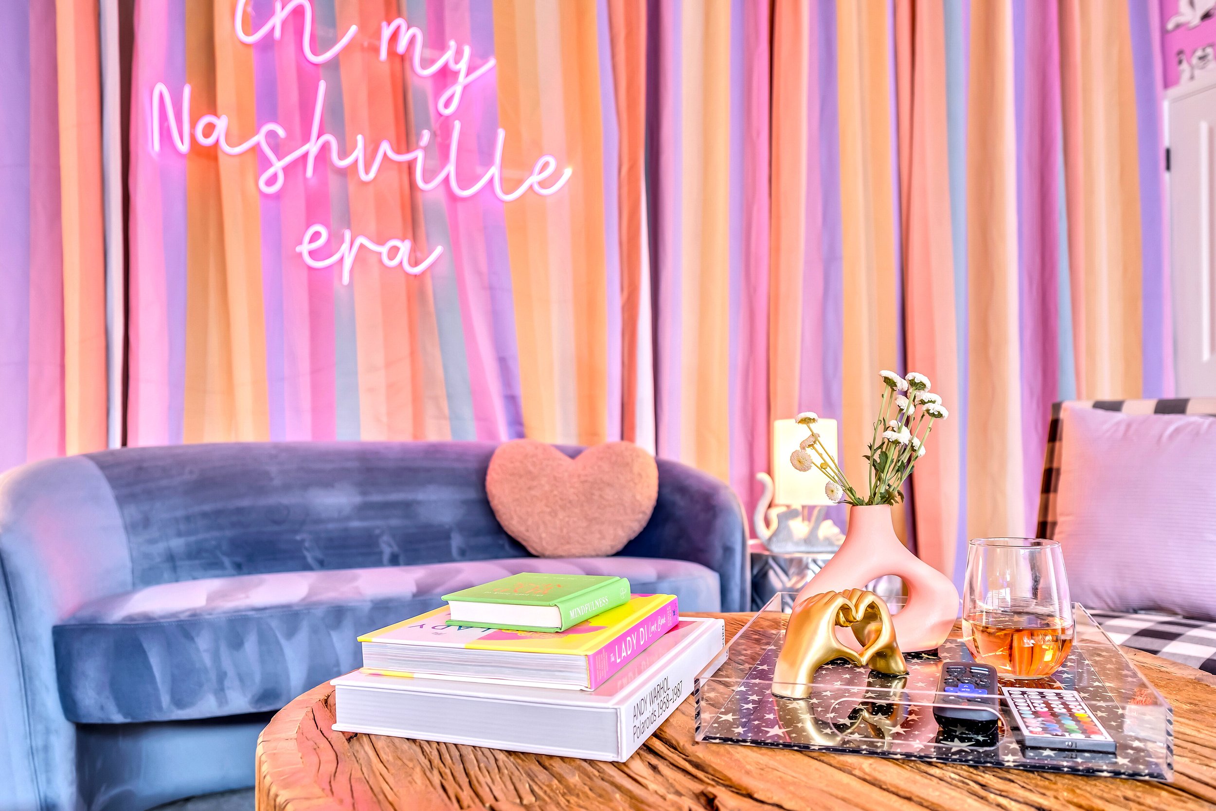 A living room with pastel-colored curtains, a neon sign saying 'Oh my NASHVILLE erA,' a blue velvet sofa with a heart-shaped pillow, a side table with a lamp and books, a flower vase, and a black and white checkered floor.