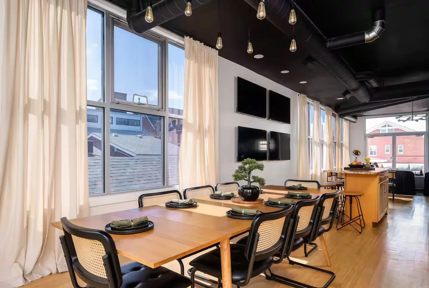 Dining area with a wooden table set with black plates and napkins, a bonsai tree centerpiece, surrounded by black and beige chairs, large windows with cream curtains, wall-mounted televisions, and a black ceiling with Edison bulbs and exposed ducts.