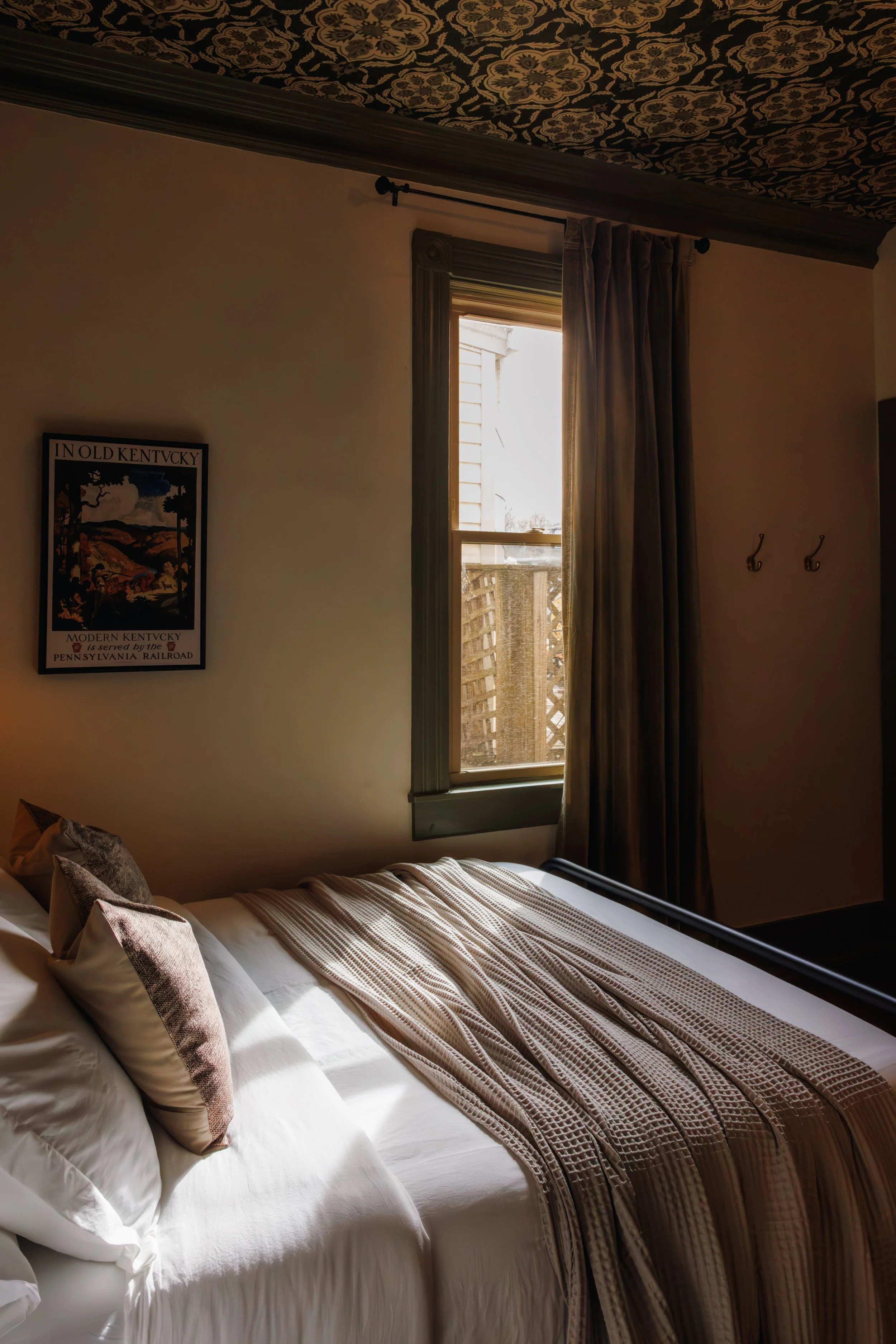 A bedroom with a bed, pillows, and a blanket. A window with curtains is above the bed, with sunlight coming through. There is a framed picture on the wall to the left of the window.