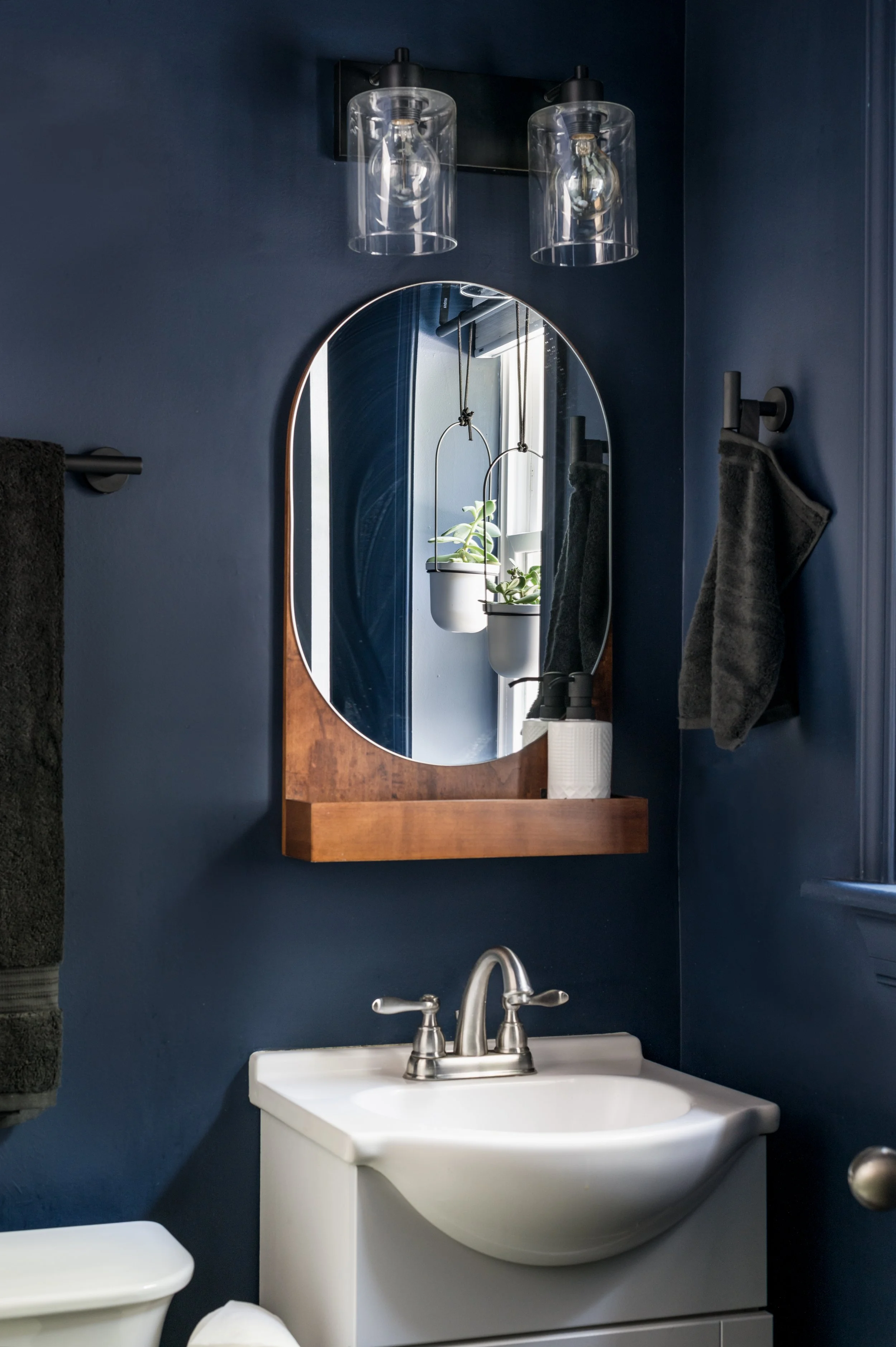 A bathroom with a dark blue wall, a white sink with a silver faucet, a mirror with a wooden frame, a small potted plant hanging in front of a window, and gray towels hanging on a black towel holder.
