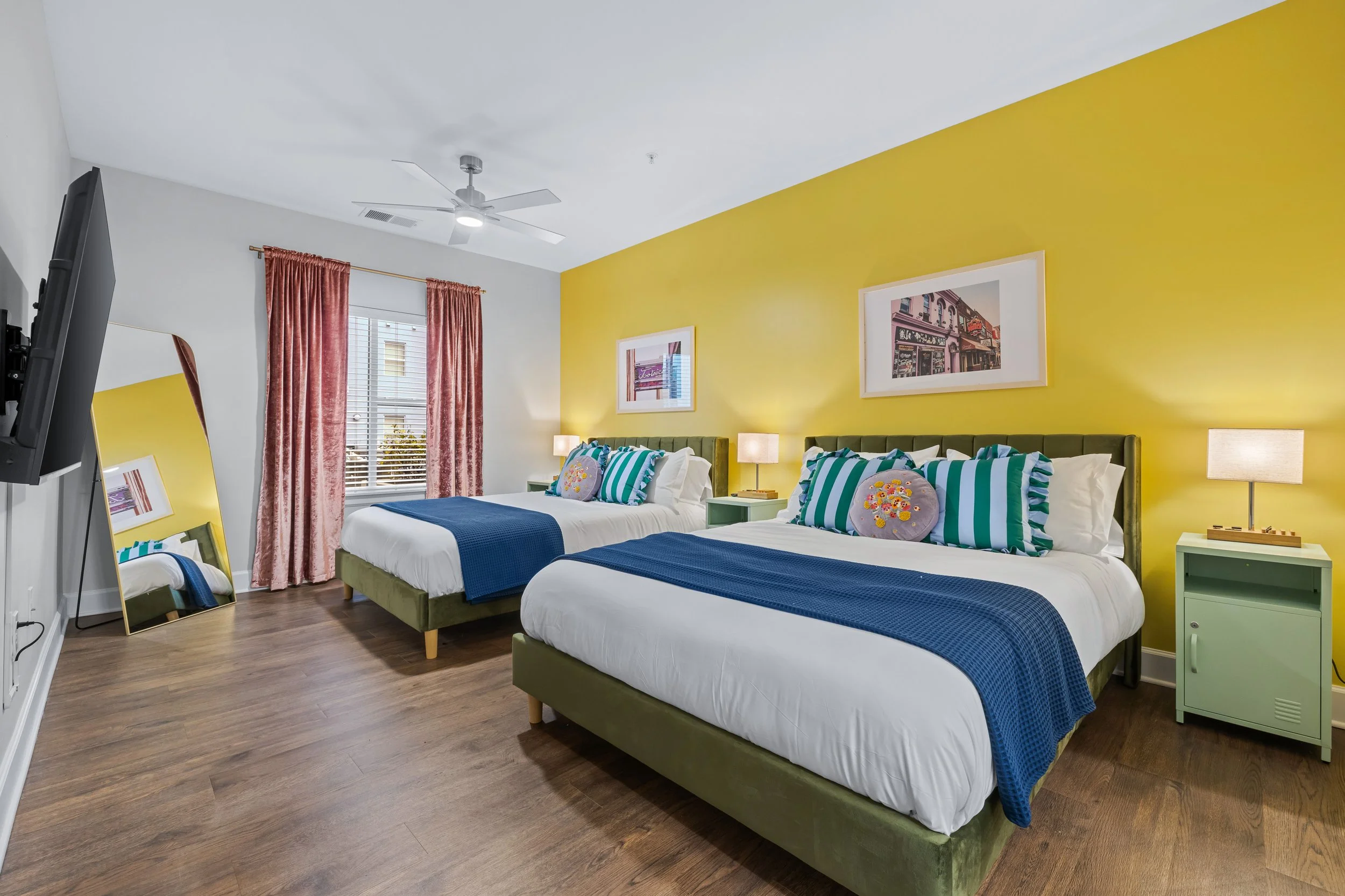 A bedroom with two beds featuring green frames and striped and floral pillows, yellow accent wall, framed pictures, pink curtains, and a window with a building view.