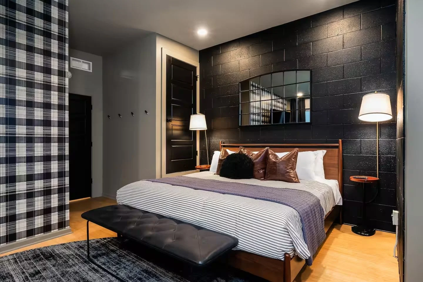 Modern bedroom with black accent wall, wooden bed frame, striped bedding, and various pillows. Features two side lamps, a reflective wall decor mirror, a black door, and a patterned area rug.
