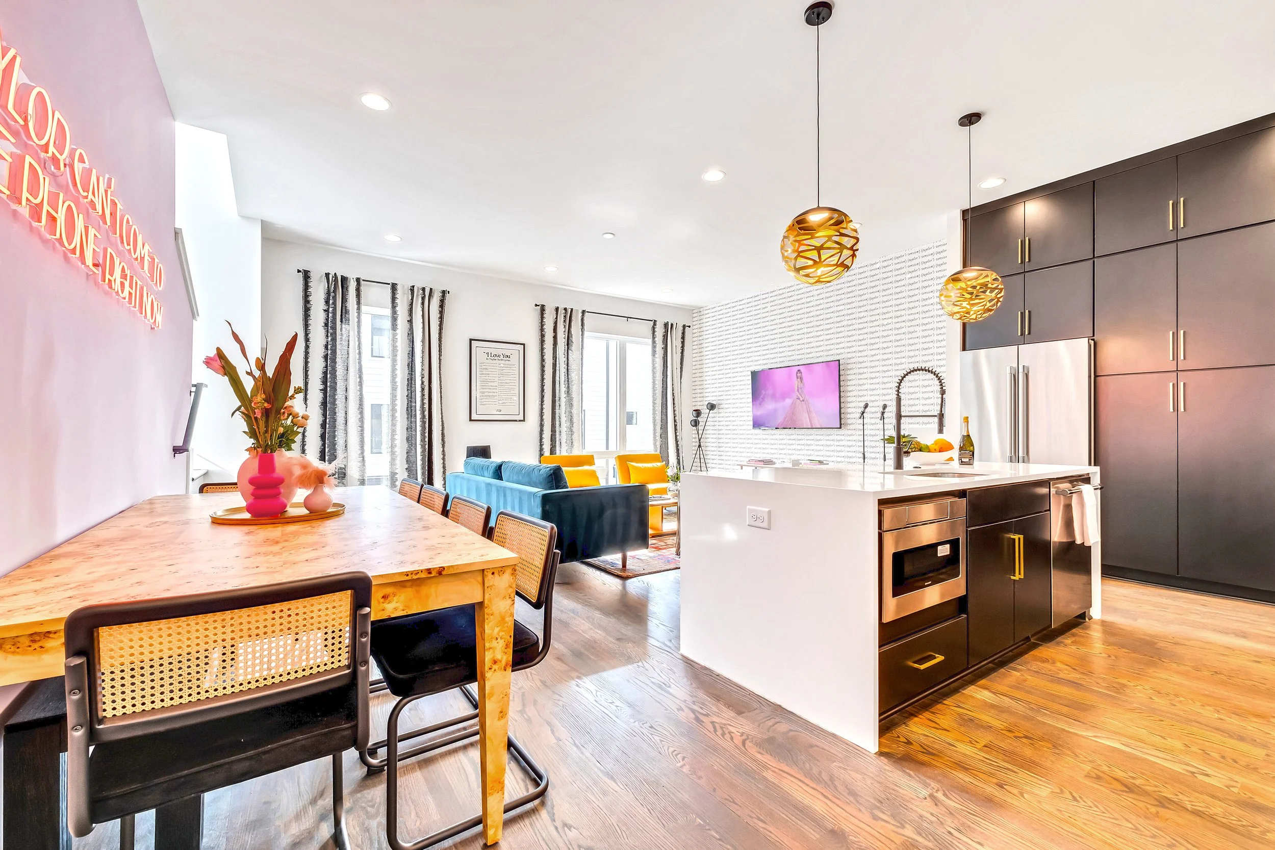 Open-concept living room and kitchen with modern decor, colorful chairs, a wooded dining table, wall art, large windows with curtains, and hanging pendant lights.
