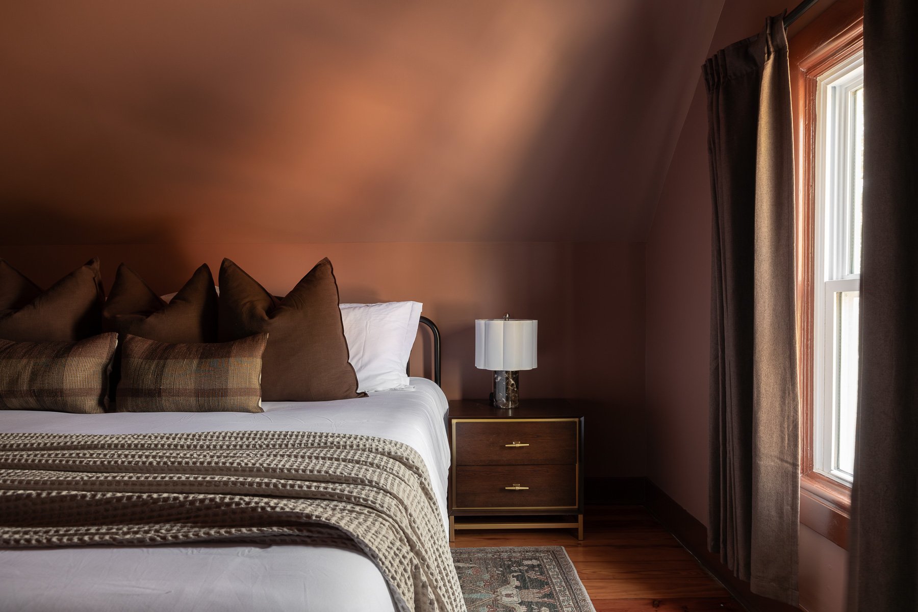 A bedroom with a neatly made bed, brown and beige pillows, a patterned blanket, a wooden nightstand with drawers, a decorative lamp, and a window with brown curtains.
