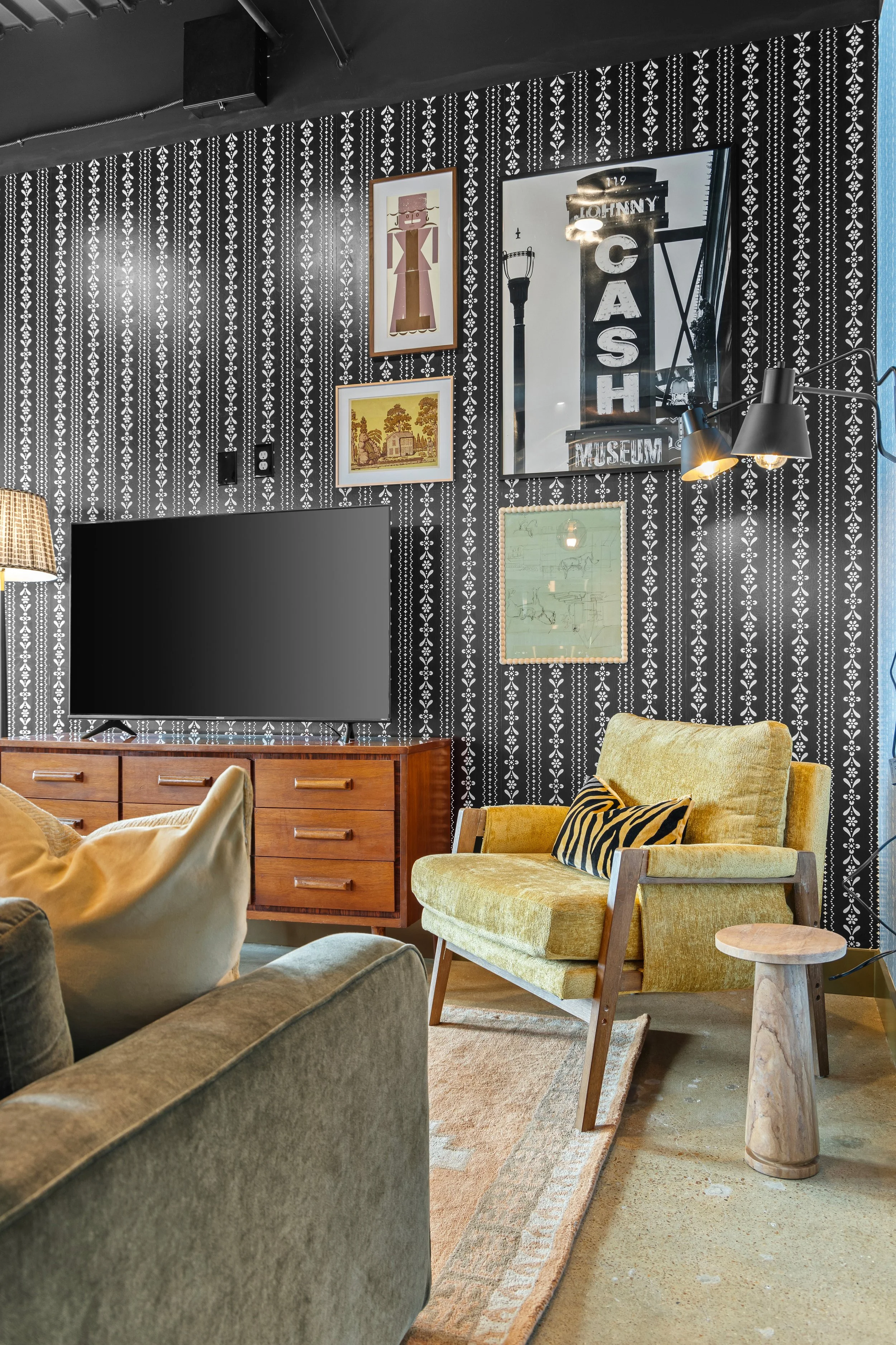 A cozy living room with a black and white patterned wall featuring framed artwork and a large mirror. There is a mustard yellow lounge chair with a black and yellow striped pillow, a wooden side table, and a vintage wooden cabinet with a TV on top. The room has a mix of modern and vintage decor with a warm, inviting atmosphere.